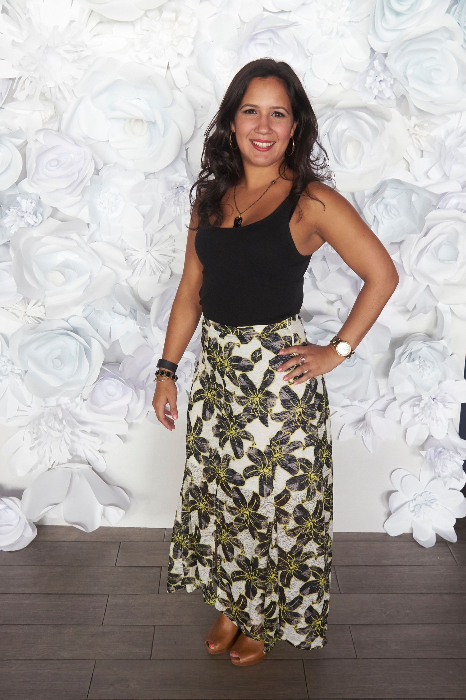 A person in a black sleeveless top and tropical print skirt poses playfully in front of a white floral backdrop.