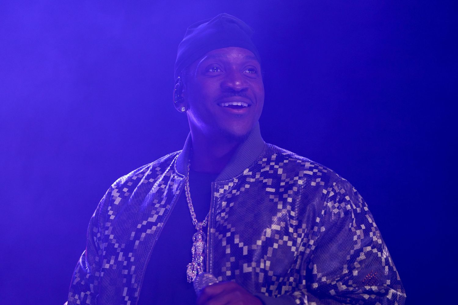 A performer wearing a patterned jacket smiles while singing on stage under dramatic blue lighting.