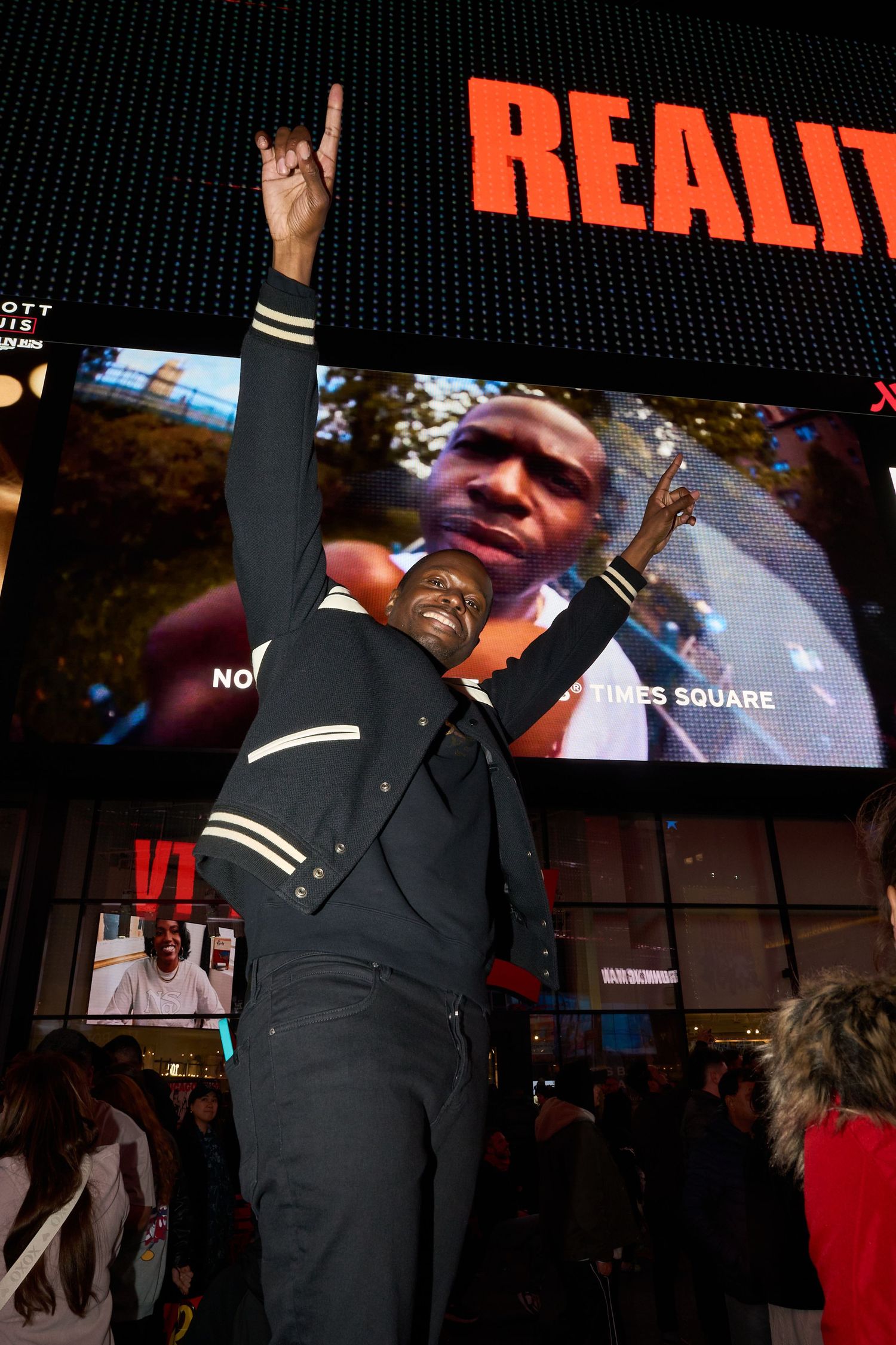 Person in black outfit raises rock hand gesture in front of large digital display in Times Square.