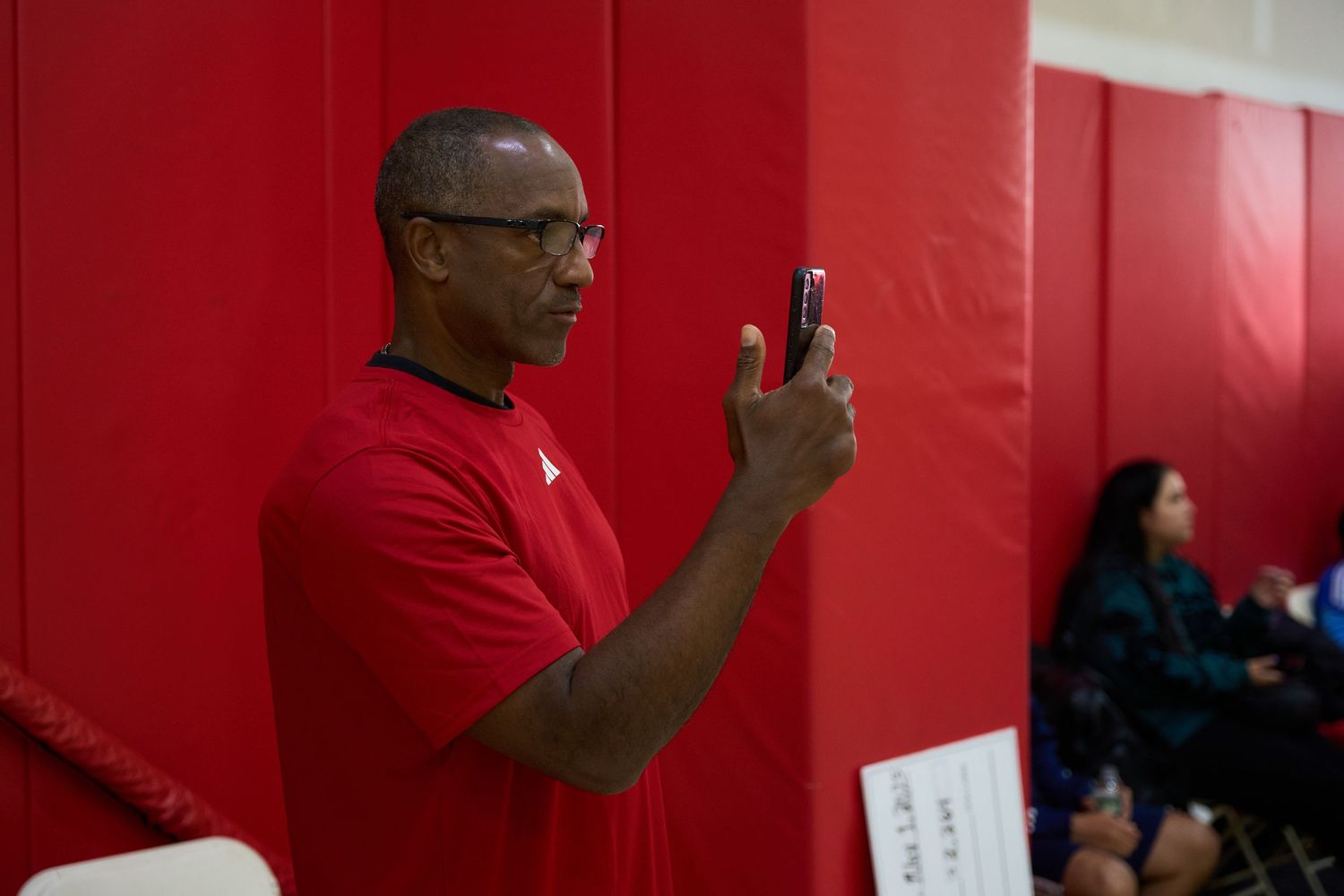 Person in red sweater taking a photo on their phone in a gymnasium with red walls.