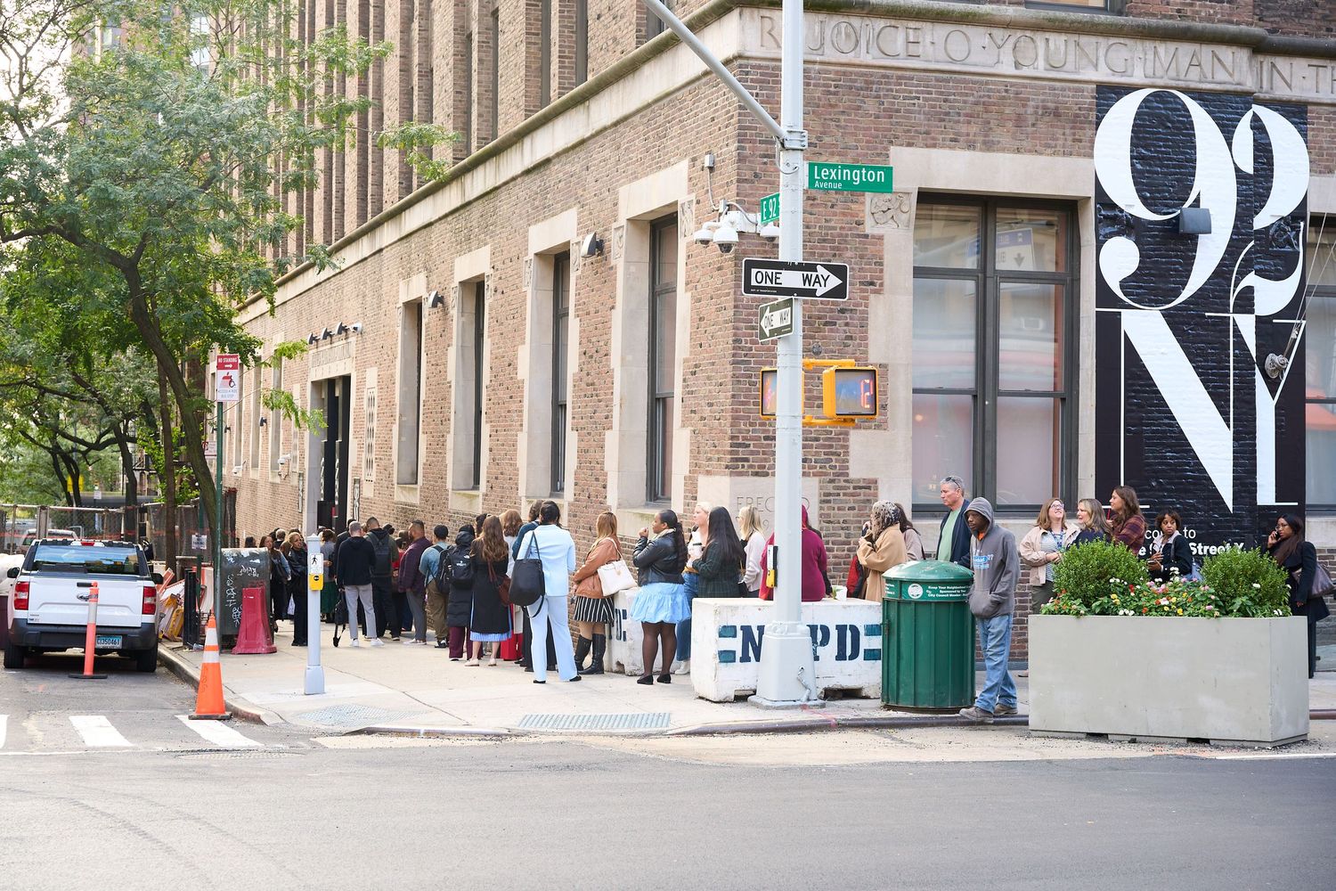 A long line of people wait outside a corner building on a city street with large numbers painted on the wall.