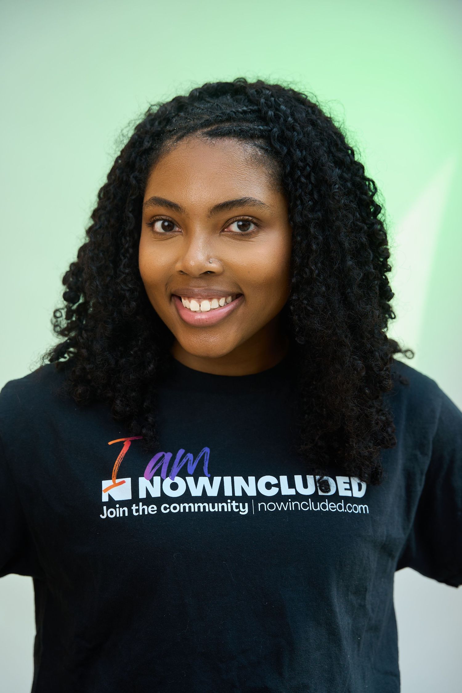 Young woman smiling in a black Now Included community t-shirt against a light green background.