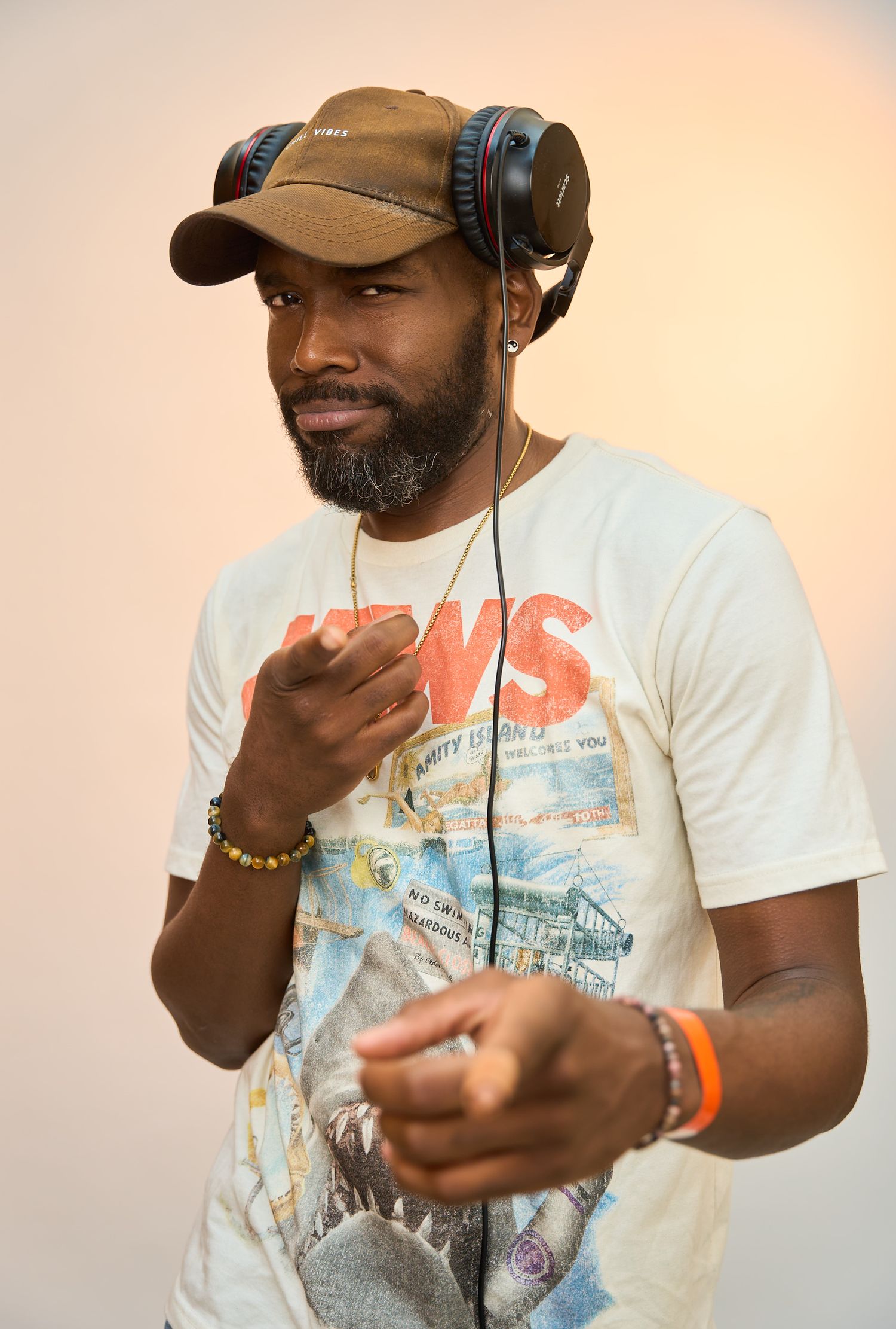 Man wearing headphones and cap gestures while DJing in casual graphic tee with beaded bracelet.