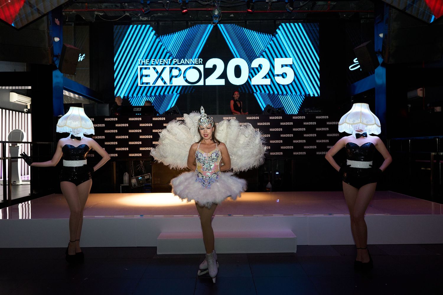 Dancers perform on stage with a bright blue EXPO2025 digital display screen in the background.