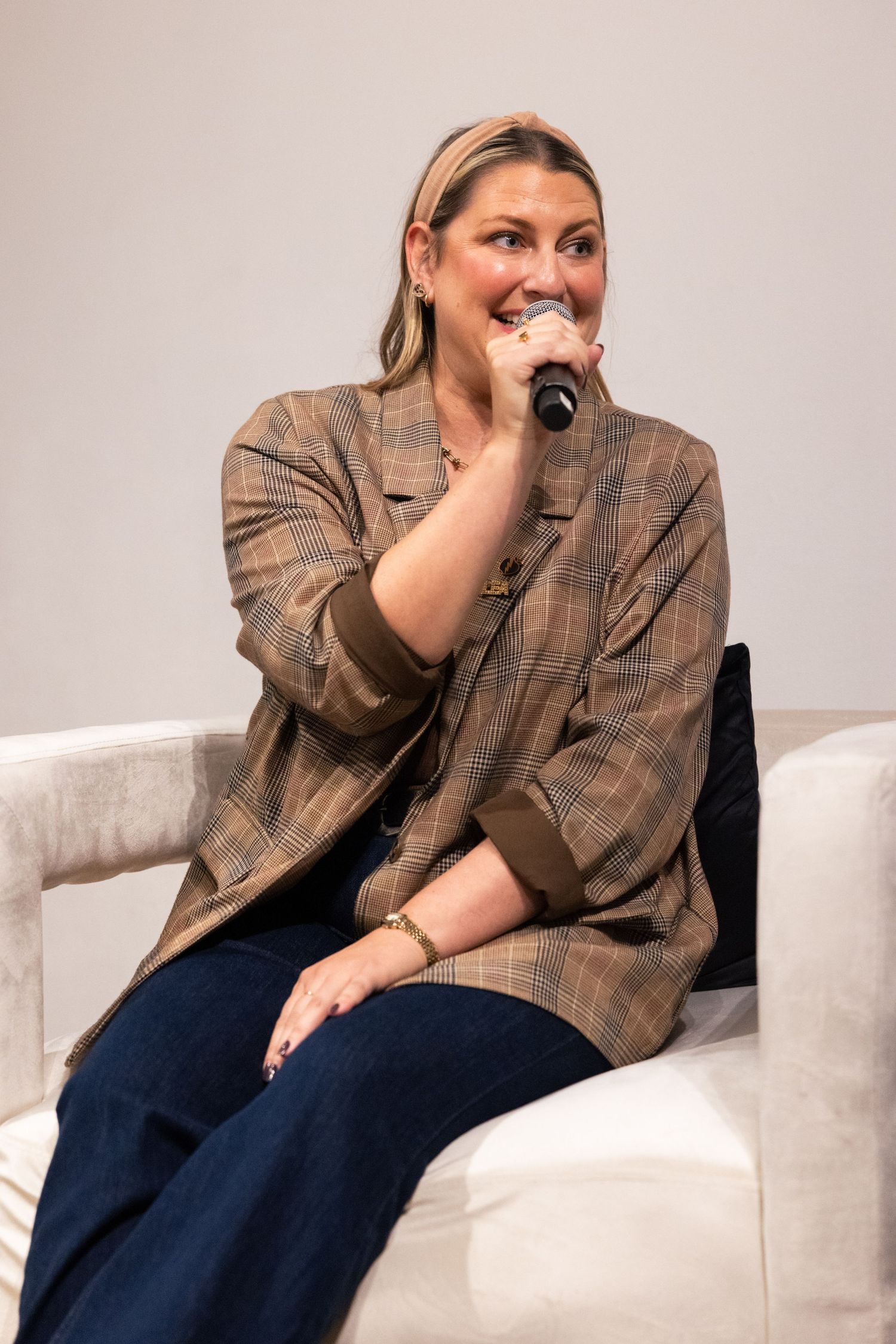 A person wearing a brown plaid blazer and dark pants sits casually on a white couch while laughing and smiling.