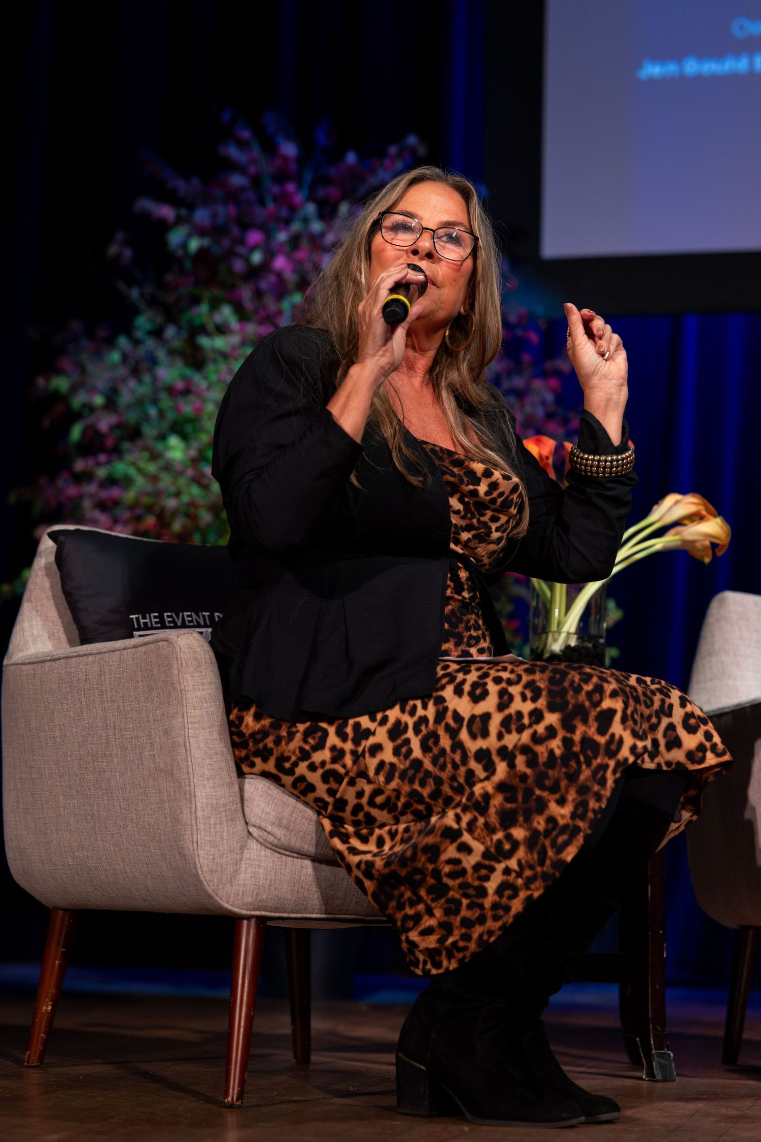 A person in a black top and leopard print pants lounges dramatically across a gray couch during a presentation.