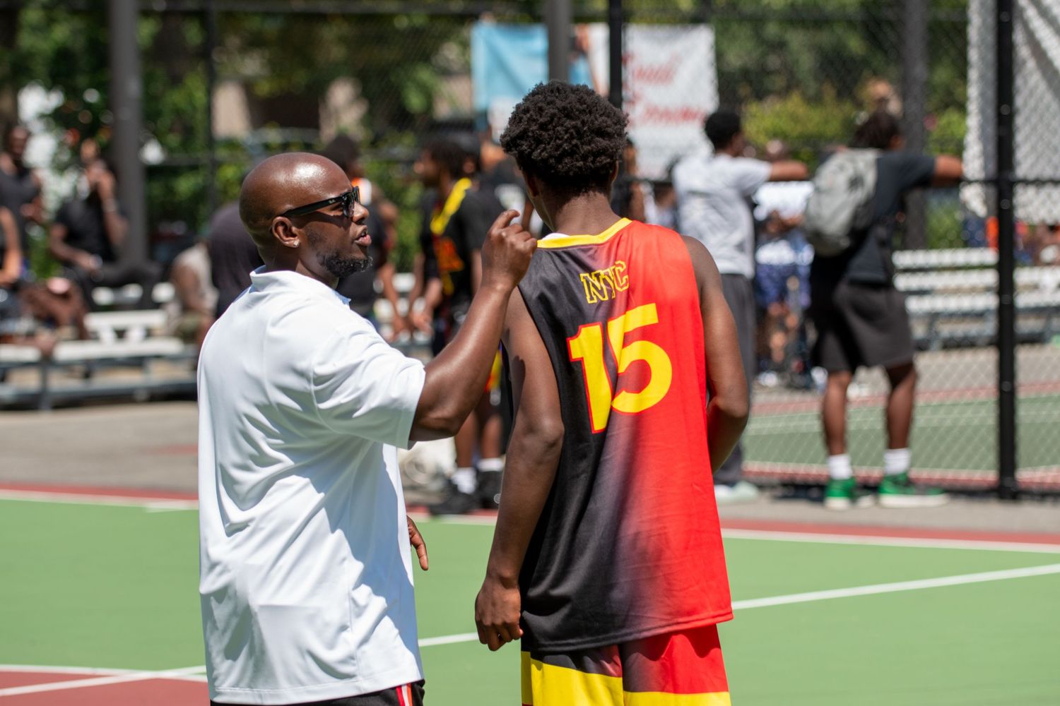 Coach gives instruction to a basketball player wearing number 15 jersey during an outdoor game on a sports court.