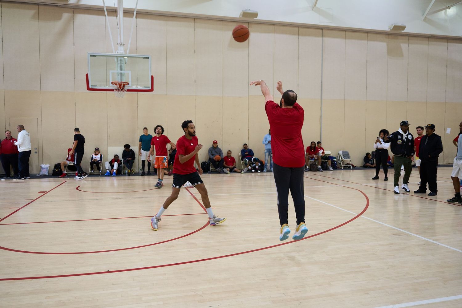 Indoor basketball game in progress with players and spectators in a gymnasium with red court markings.