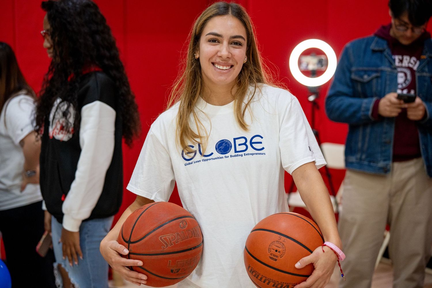 A person in a white shirt holds a basketball in a gym setting with red and blue accents visible in the background.
