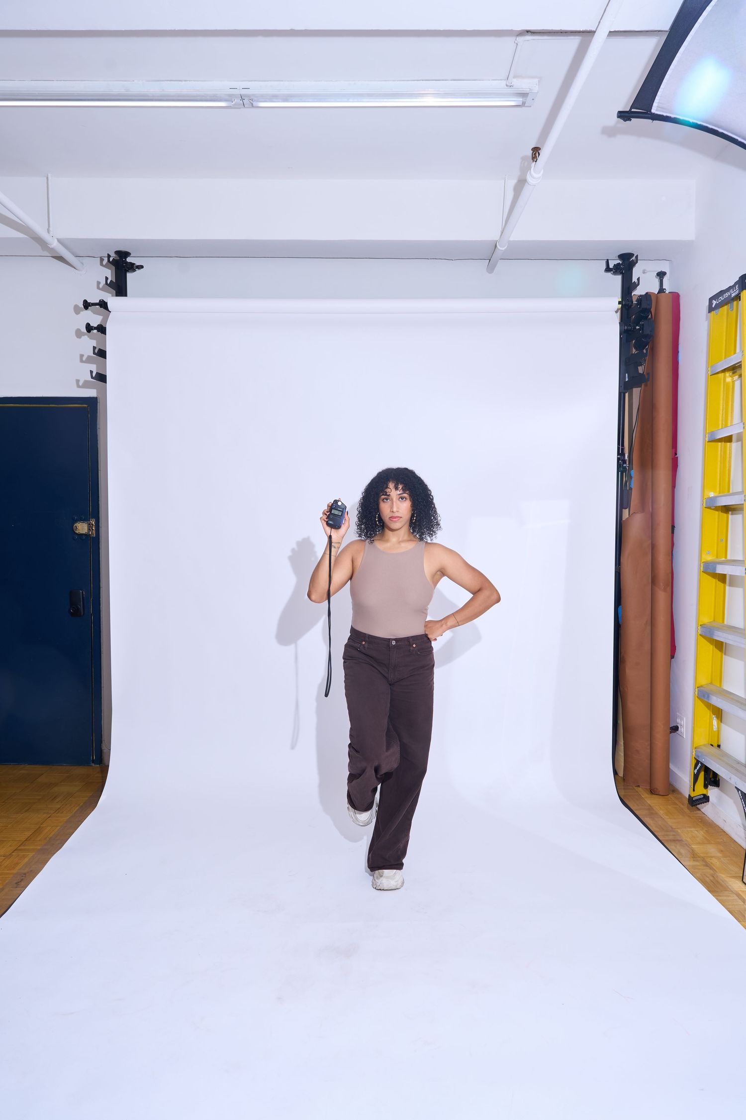 Person in beige top and brown pants holding a camera in a professional white backdrop photography studio.