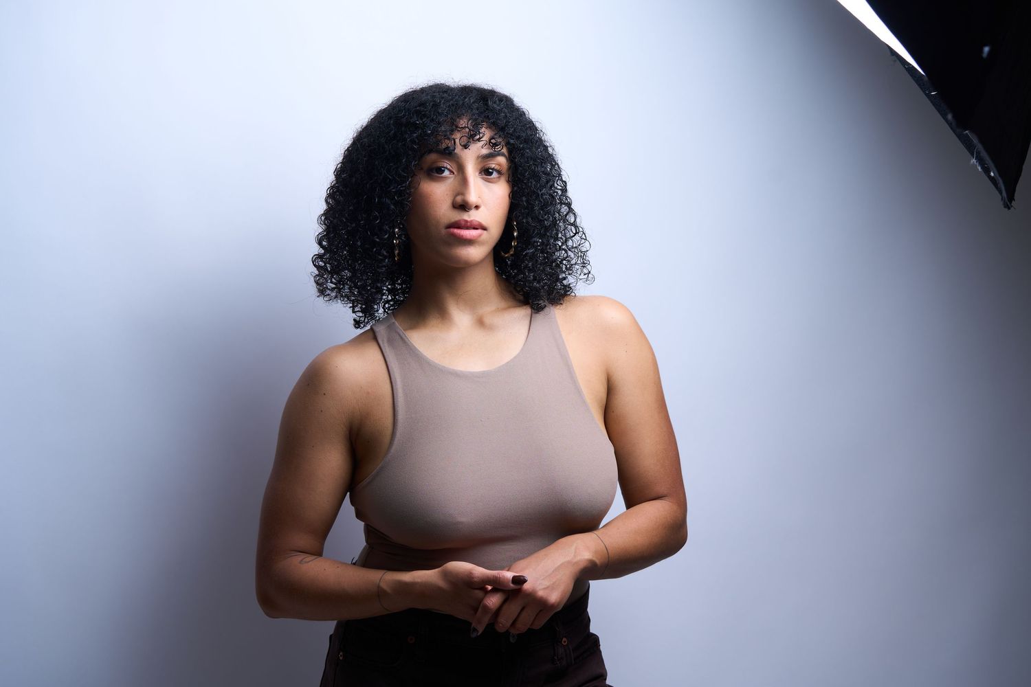 Young woman with curly hair poses confidently in a beige bodysuit against a white studio backdrop.