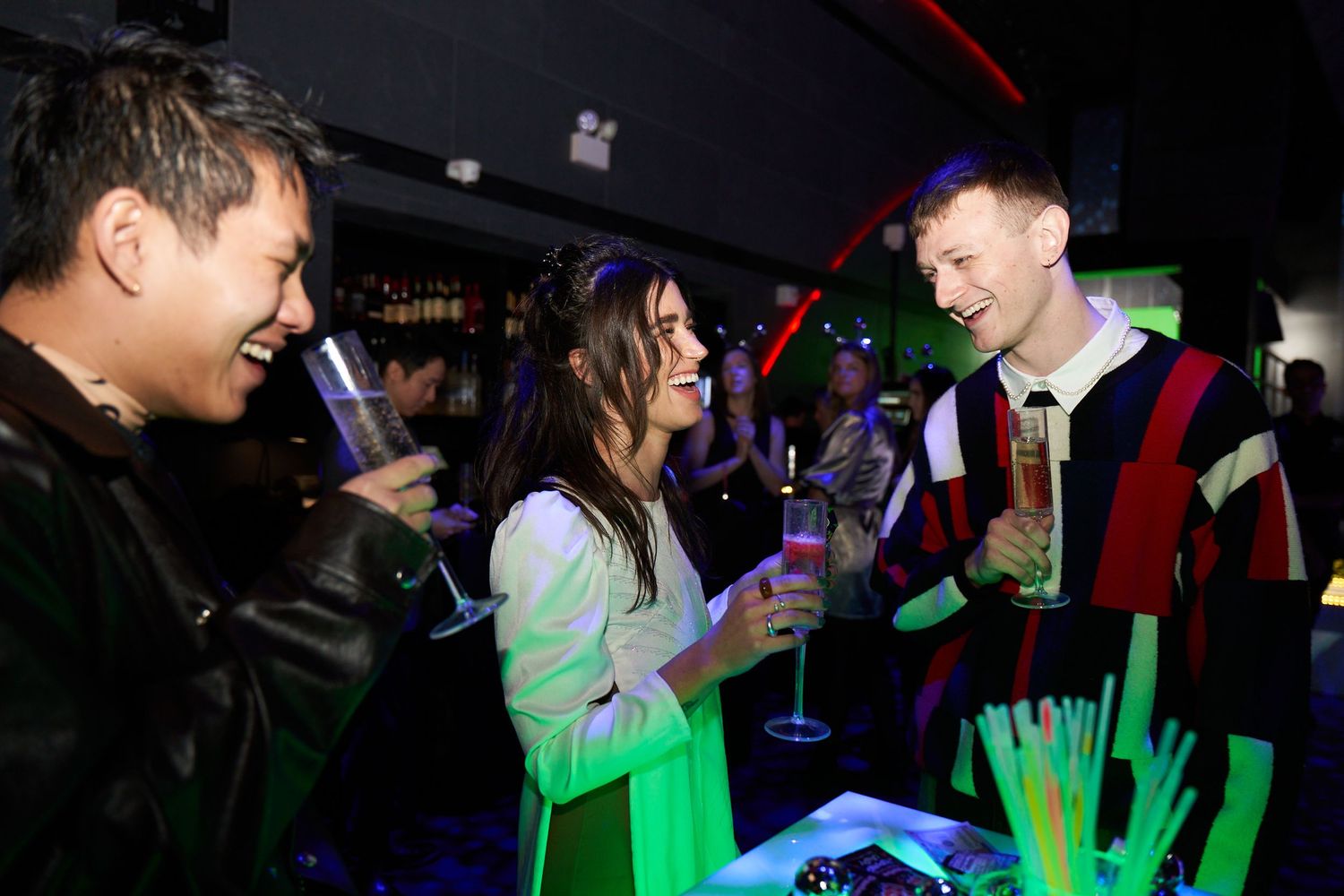People enjoying drinks and conversation at a nightclub with neon lighting and festive atmosphere.