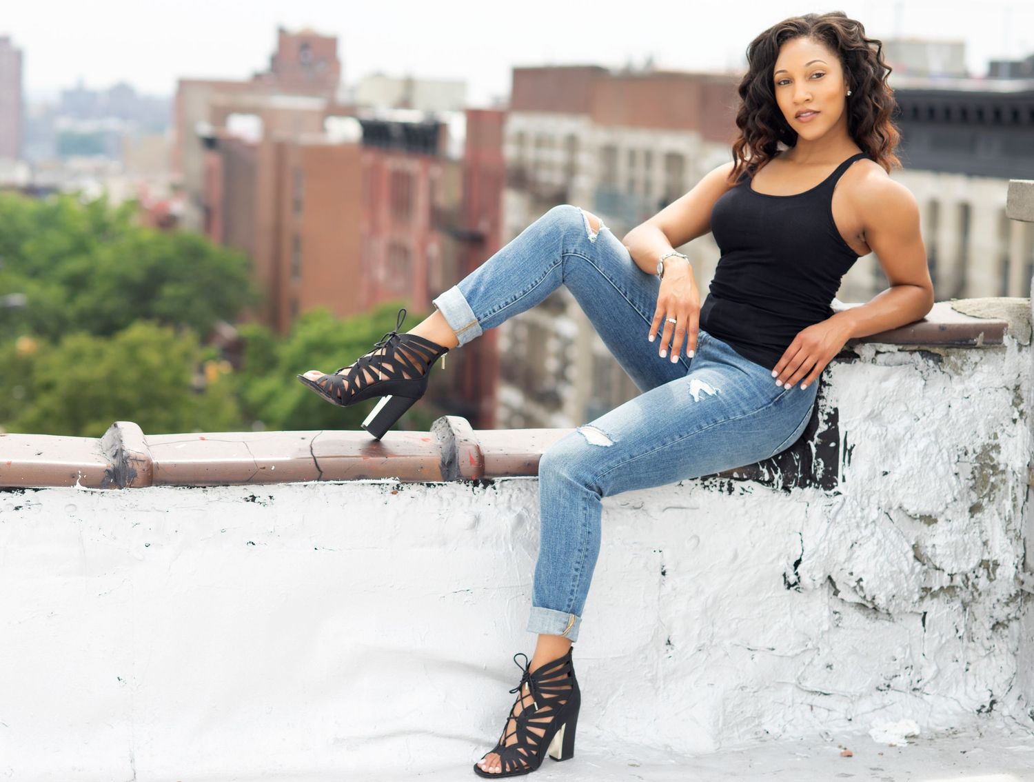 A person in a black tank top and distressed jeans sits casually on a rooftop ledge overlooking city buildings.