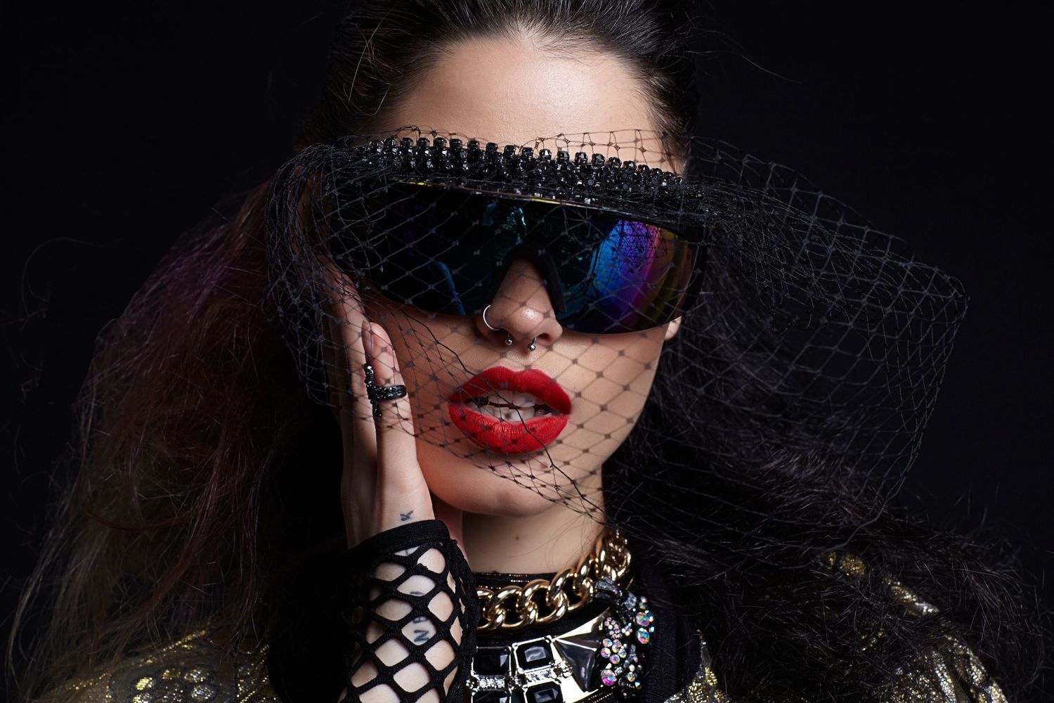 Dramatic fashion portrait with black lace veil, reflective sunglasses, red lipstick and statement jewelry against dark background.