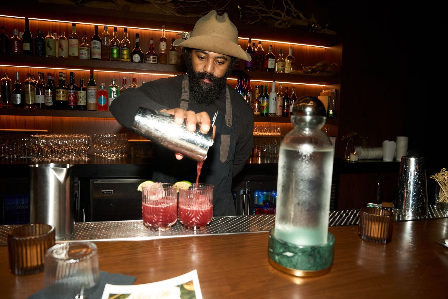Bearded bartender in a hat pours a pink cocktail from a shaker into two garnished glasses at a dimly lit bar with a bottle of water.