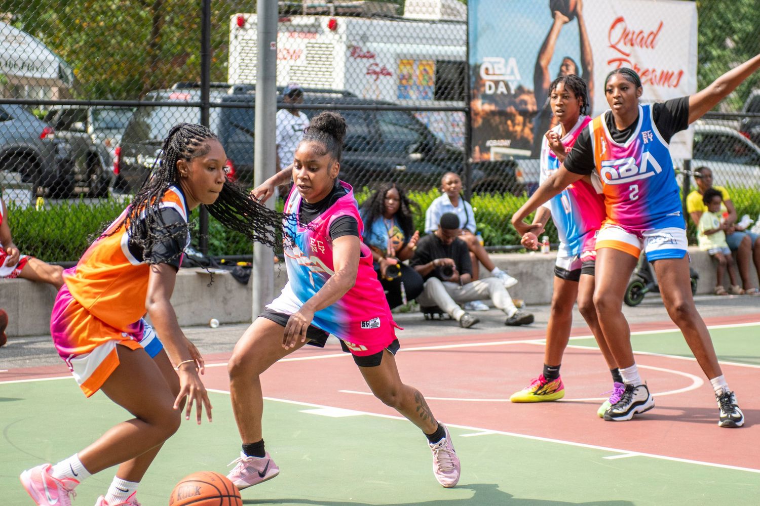 Basketball players in colorful uniforms compete aggressively on an outdoor court during a summer game.