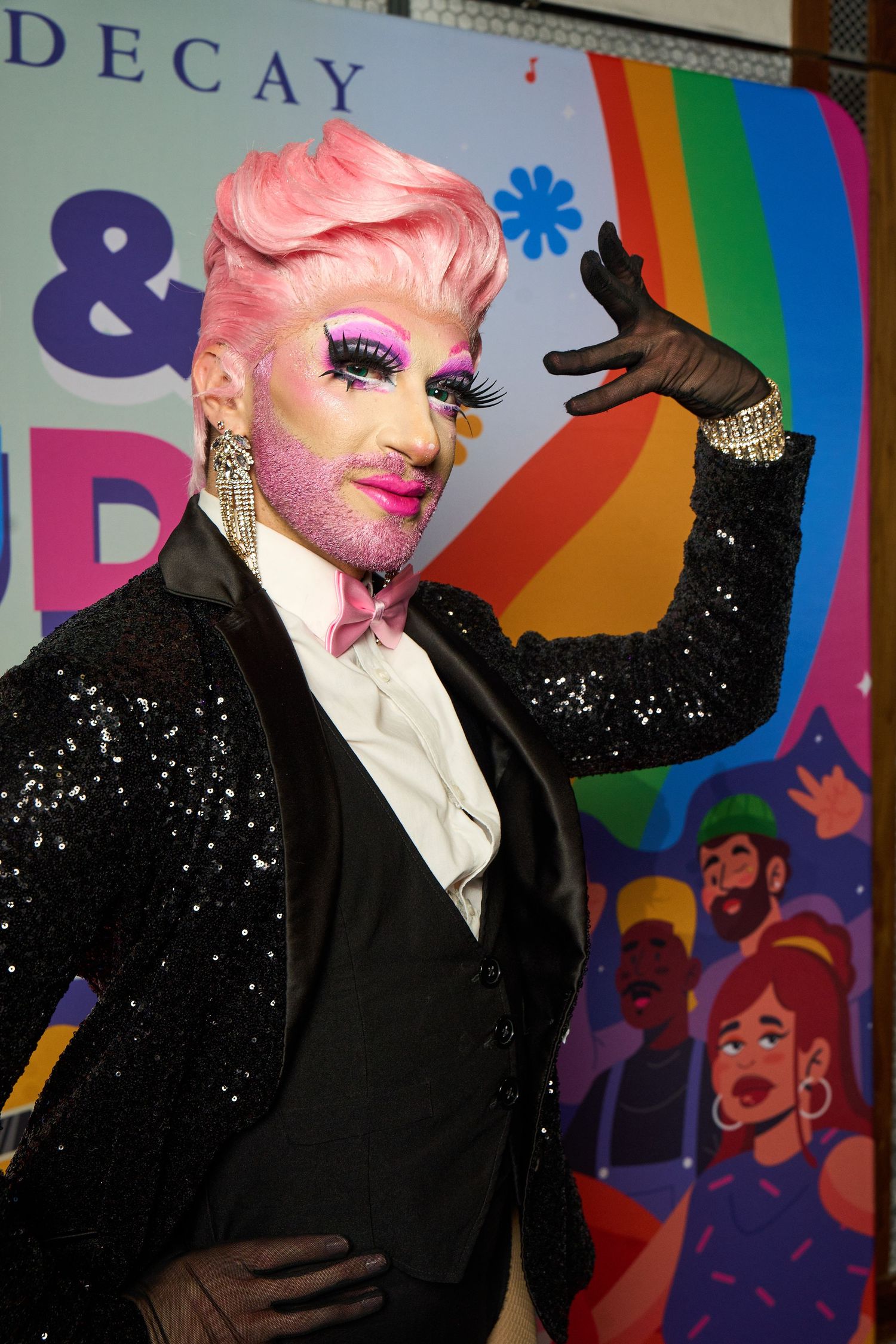 A performer in glamorous drag attire with pink hair and a sparkly black blazer poses against a colorful rainbow backdrop.