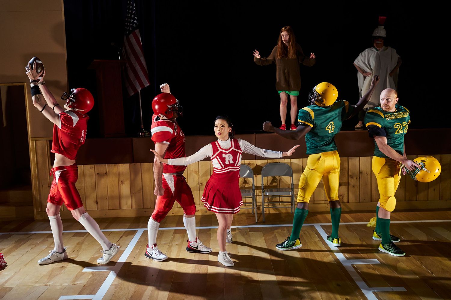 School performers in red and green costumes strike dynamic poses during a high school musical performance on stage.