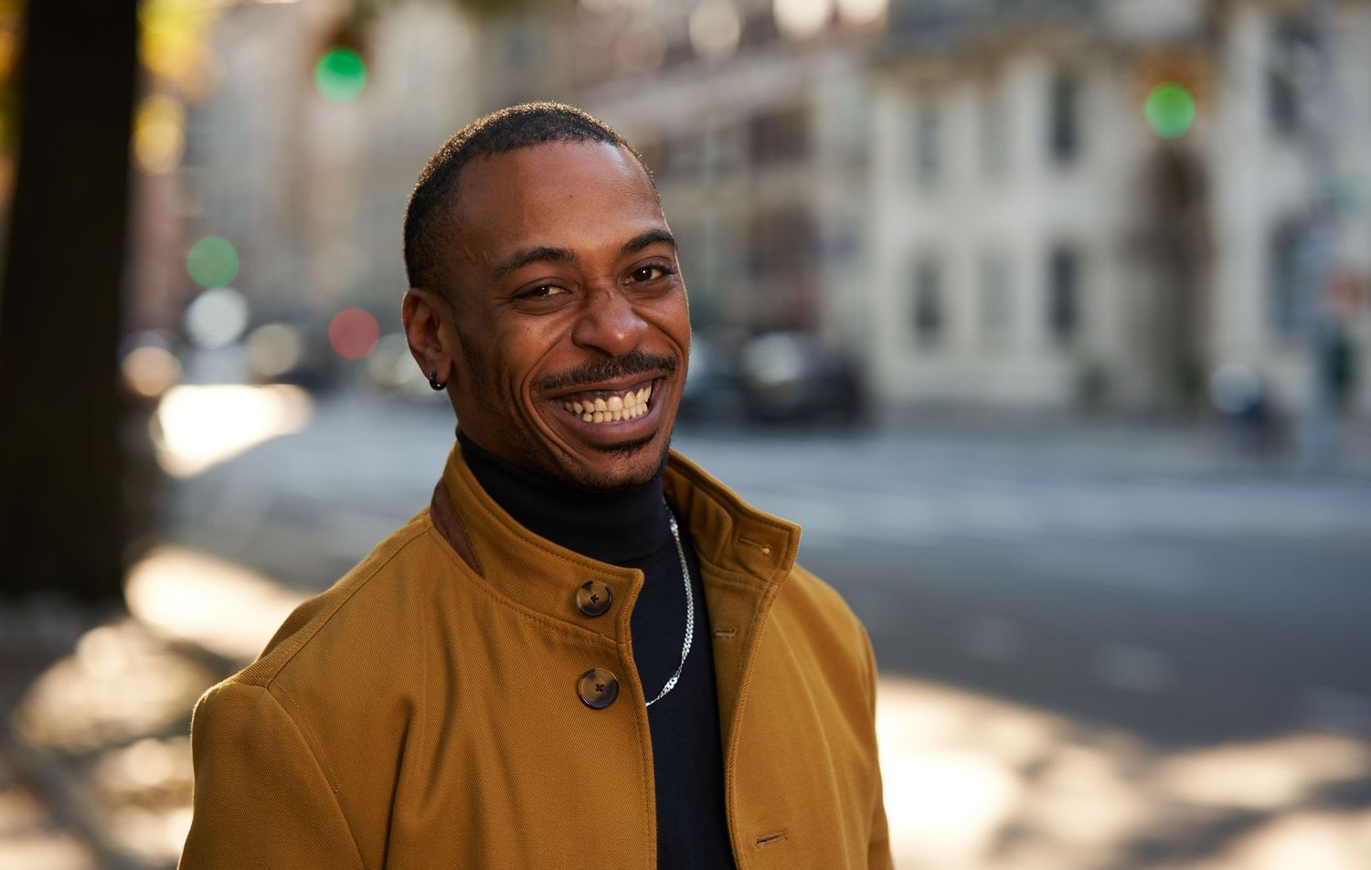 Someone wearing a brown jacket smiles while standing outdoors on a sunny day.