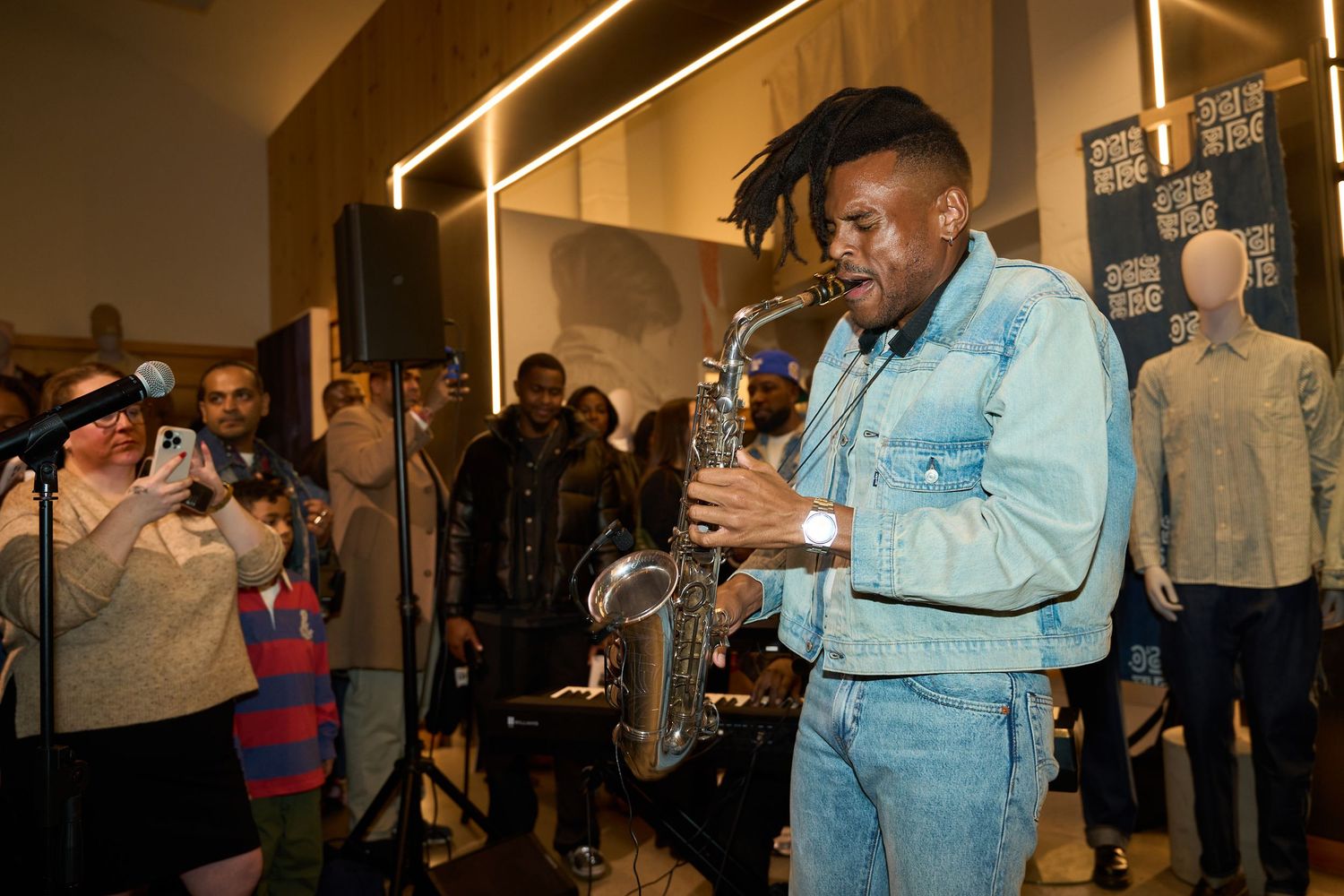 A performer in a light denim outfit sings into a microphone at an indoor venue while audience members watch in the background.