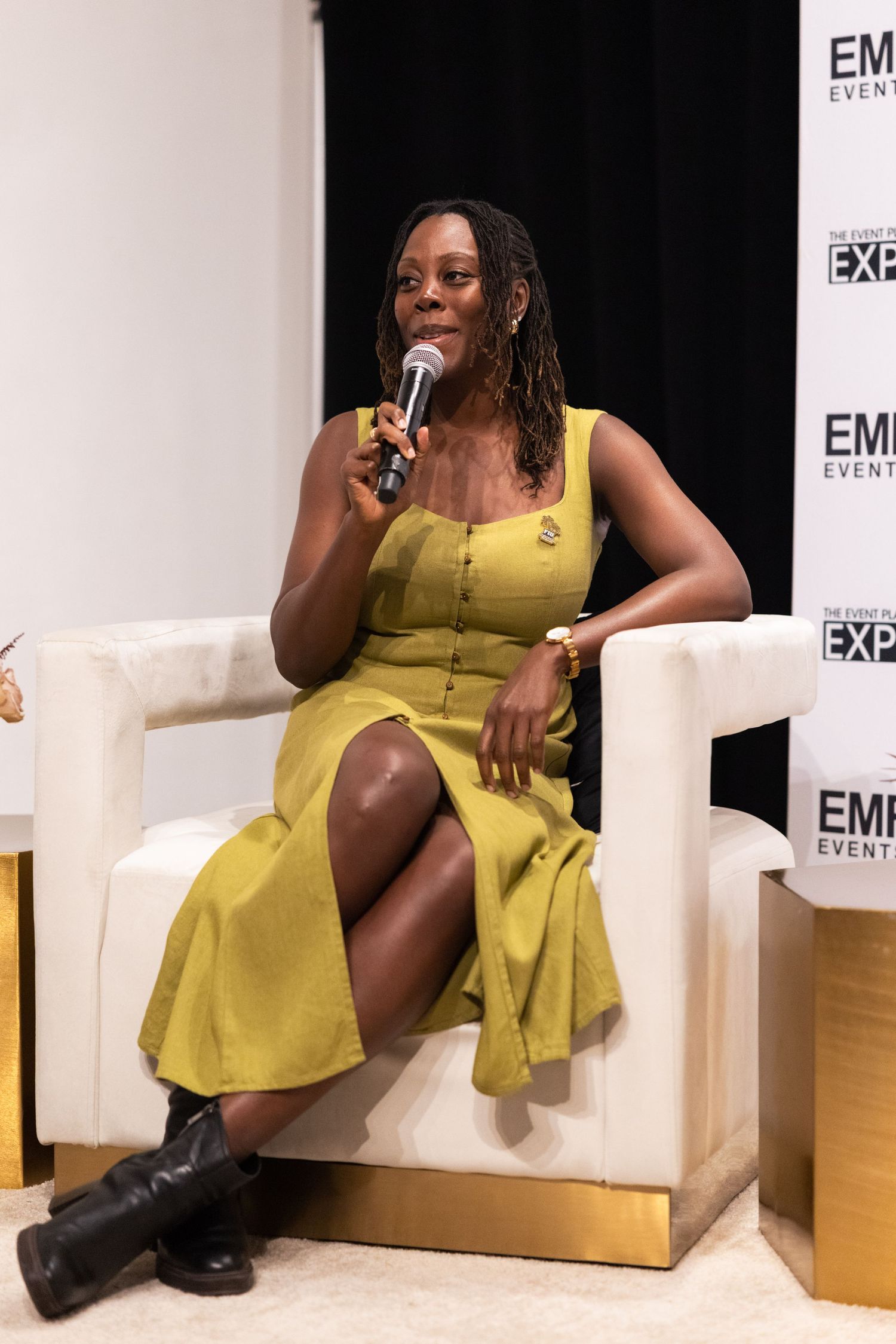 A person in a yellow dress sits in a white chair at an event, with 'EM' signage visible in the background.