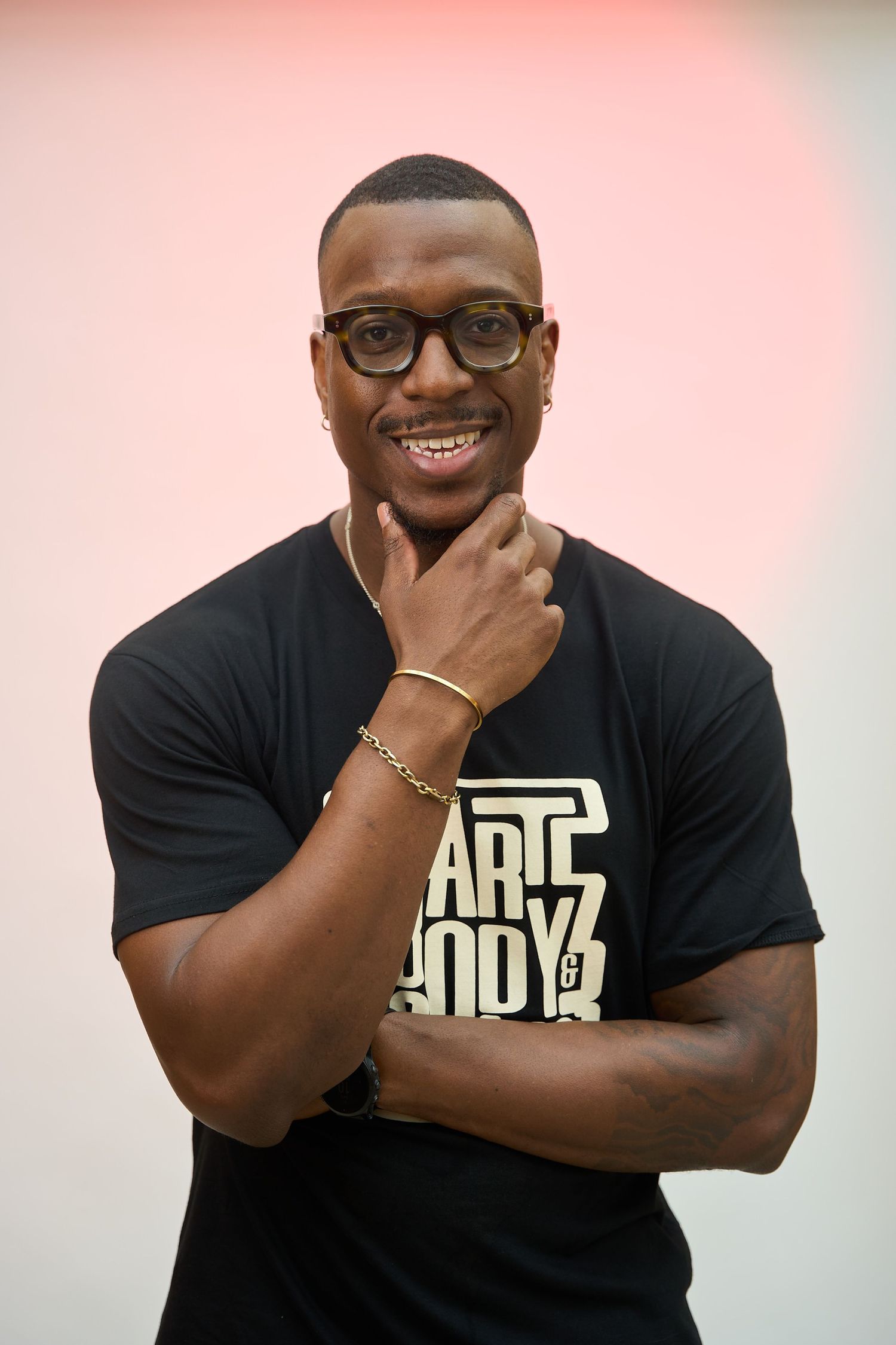 Smiling young Black man in black graphic t-shirt posing with hand on chin against a soft pink background.