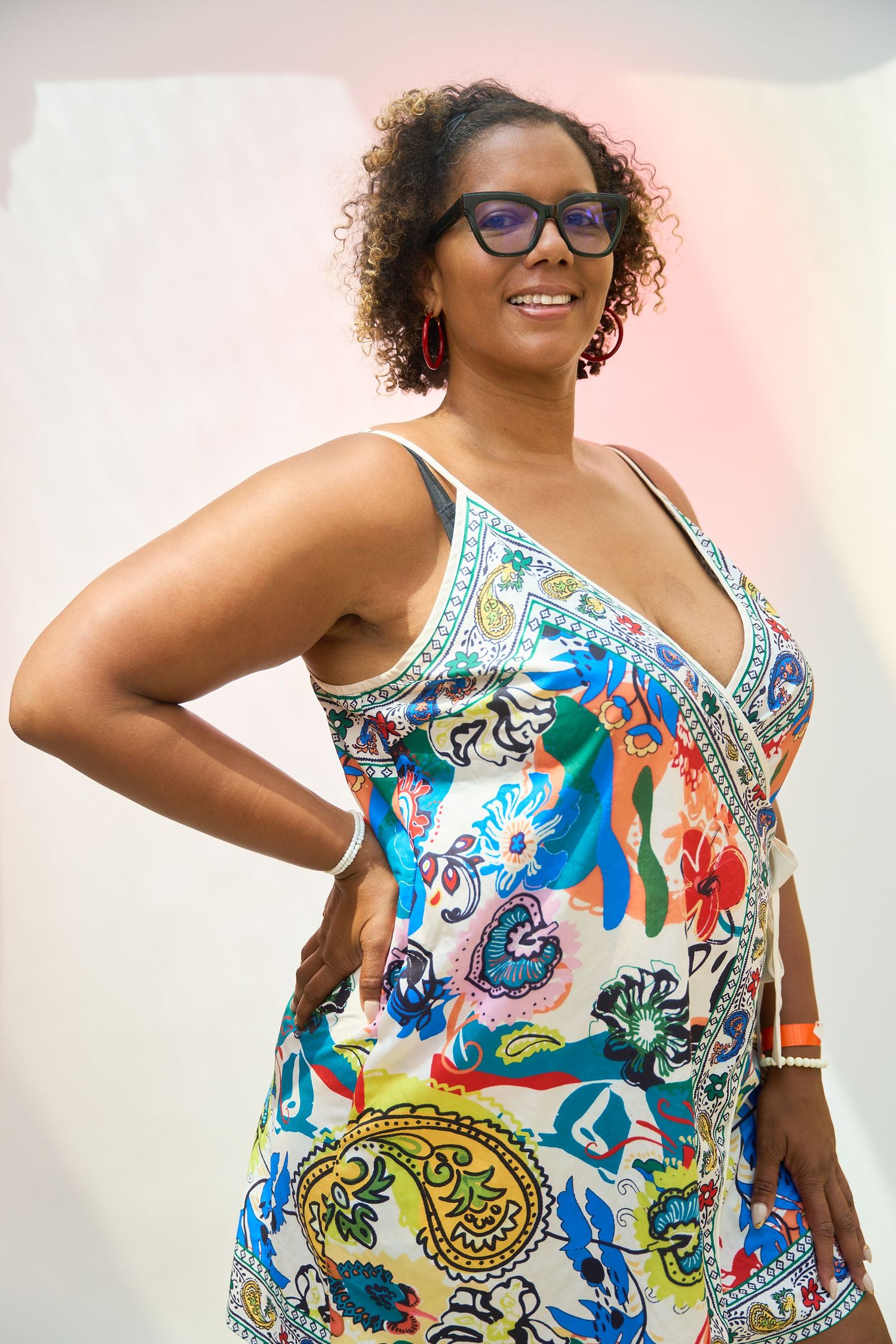 Smiling woman in colorful floral dress and glasses posing confidently with hand on hip against pastel background.
