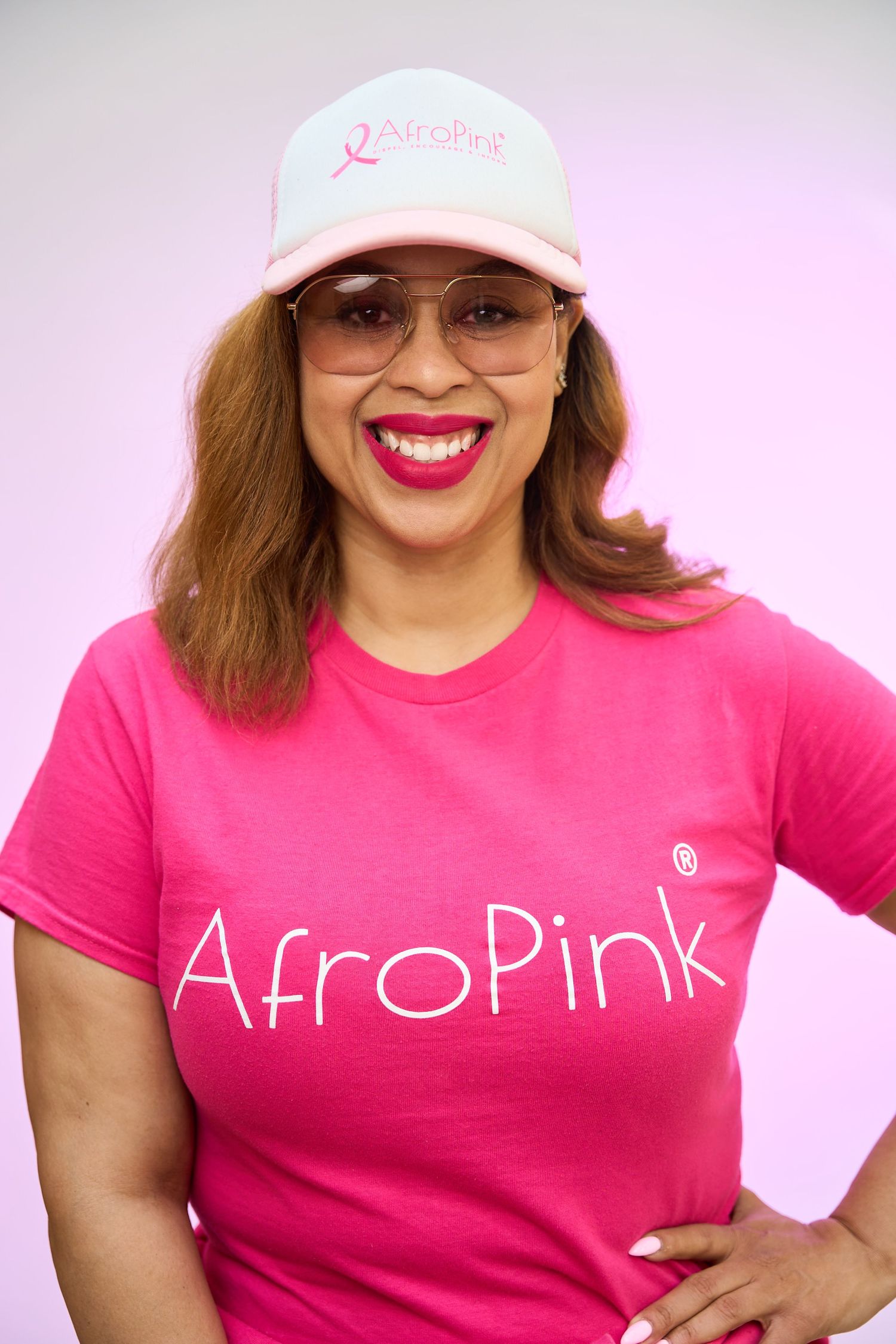 Smiling woman wearing a pink AfroPink t-shirt and matching cap, posing confidently against a light purple background.