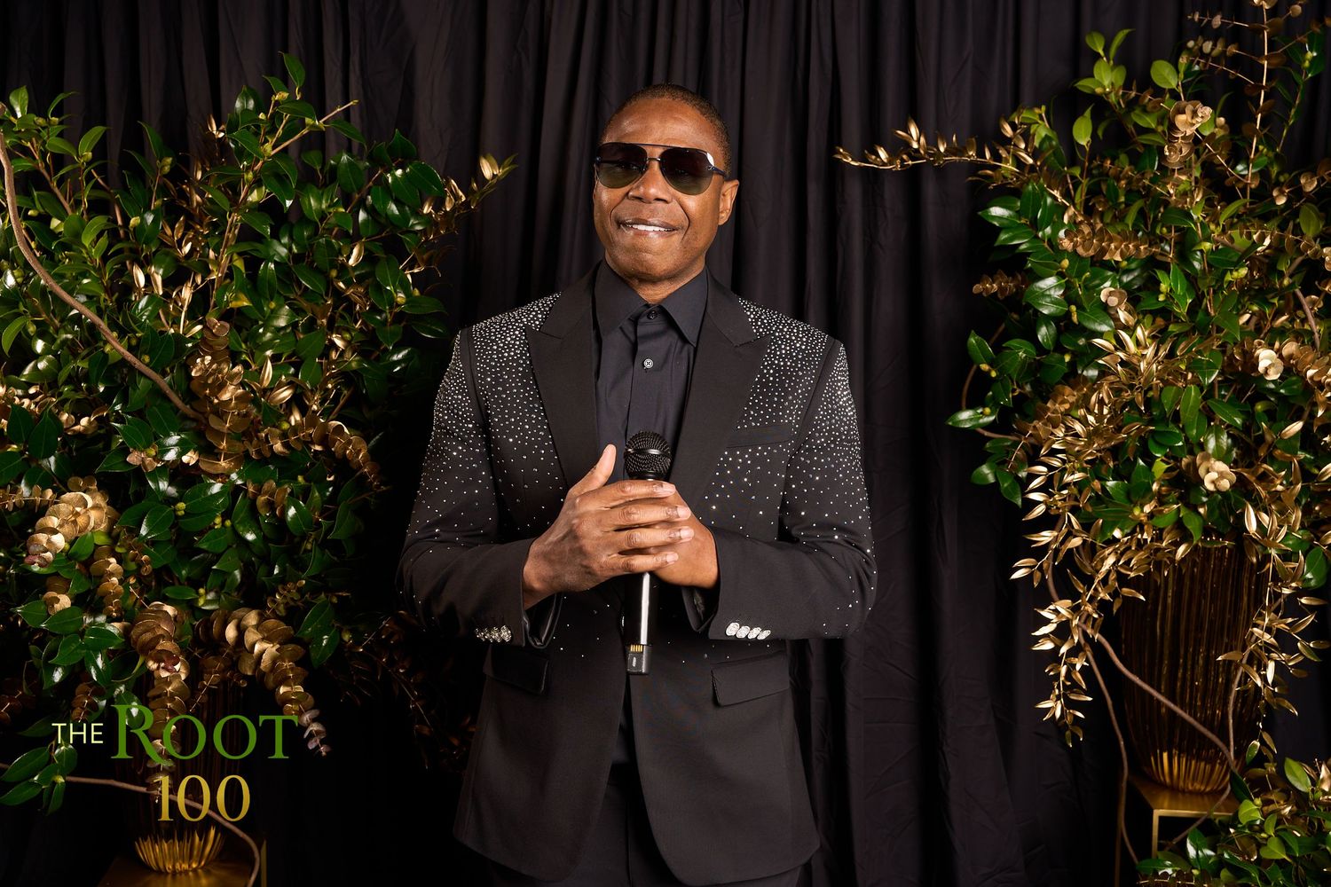 A person in a black suit and sunglasses stands in front of a dark curtain with decorative gold-lit foliage.