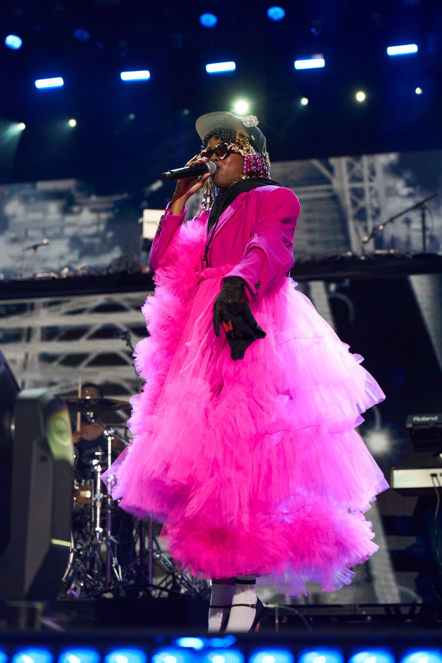 Performer in bright pink suit and tulle dress sings on stage with dramatic blue stage lighting in the background.