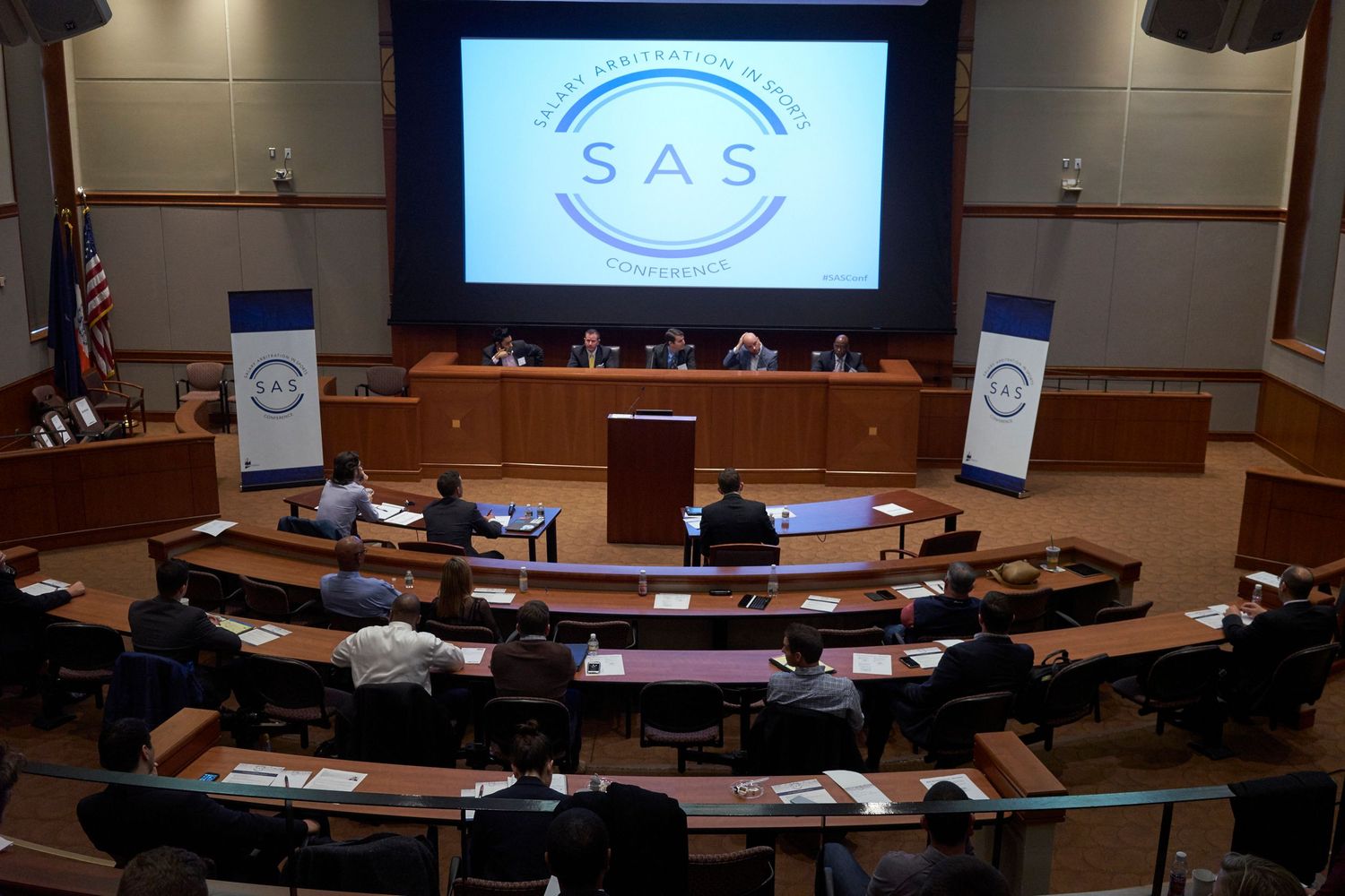 People attend a government meeting in a semicircular auditorium with SAS logo displayed on the projection screen.