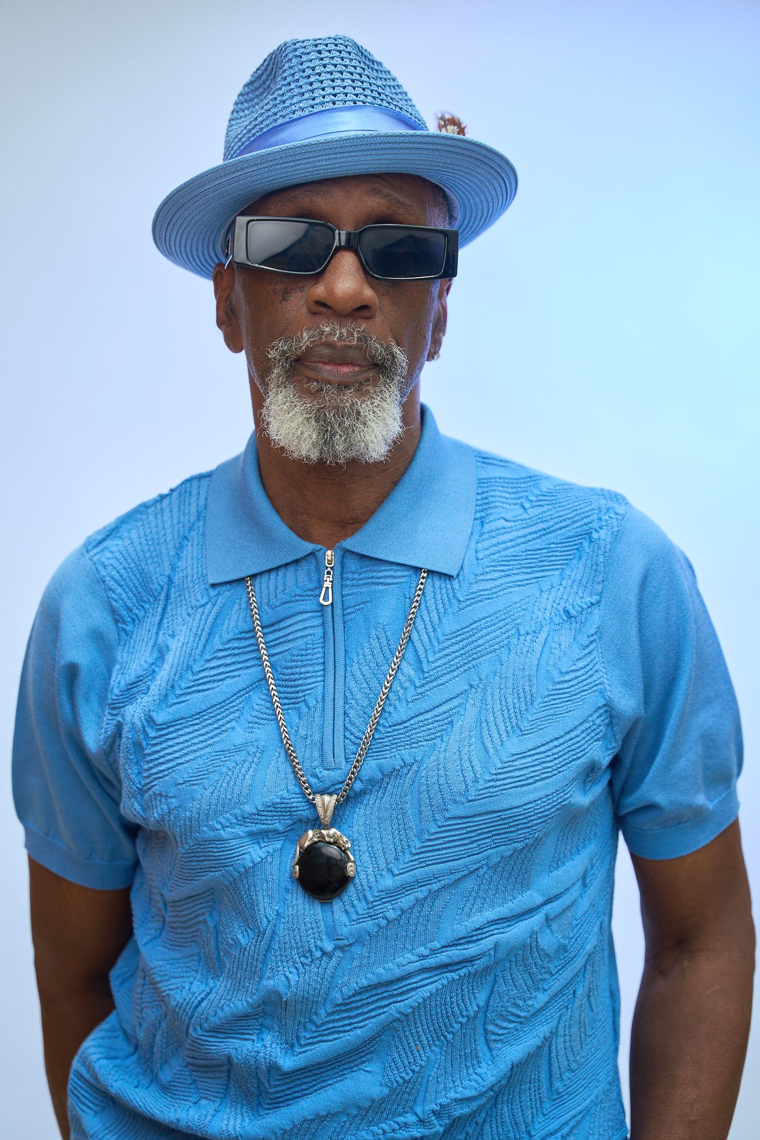 Stylish older man in blue textured polo, matching fedora, sunglasses, and chain necklace posing confidently.