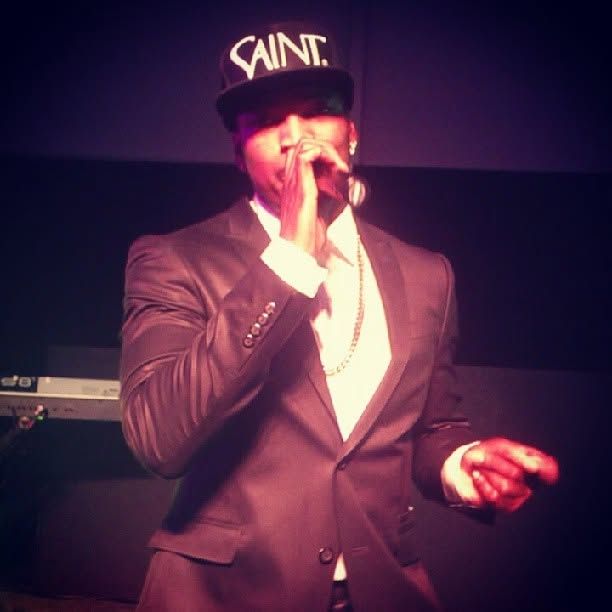 Performer in a dark suit and black SAINT cap singing into a microphone on stage under warm purple lighting.