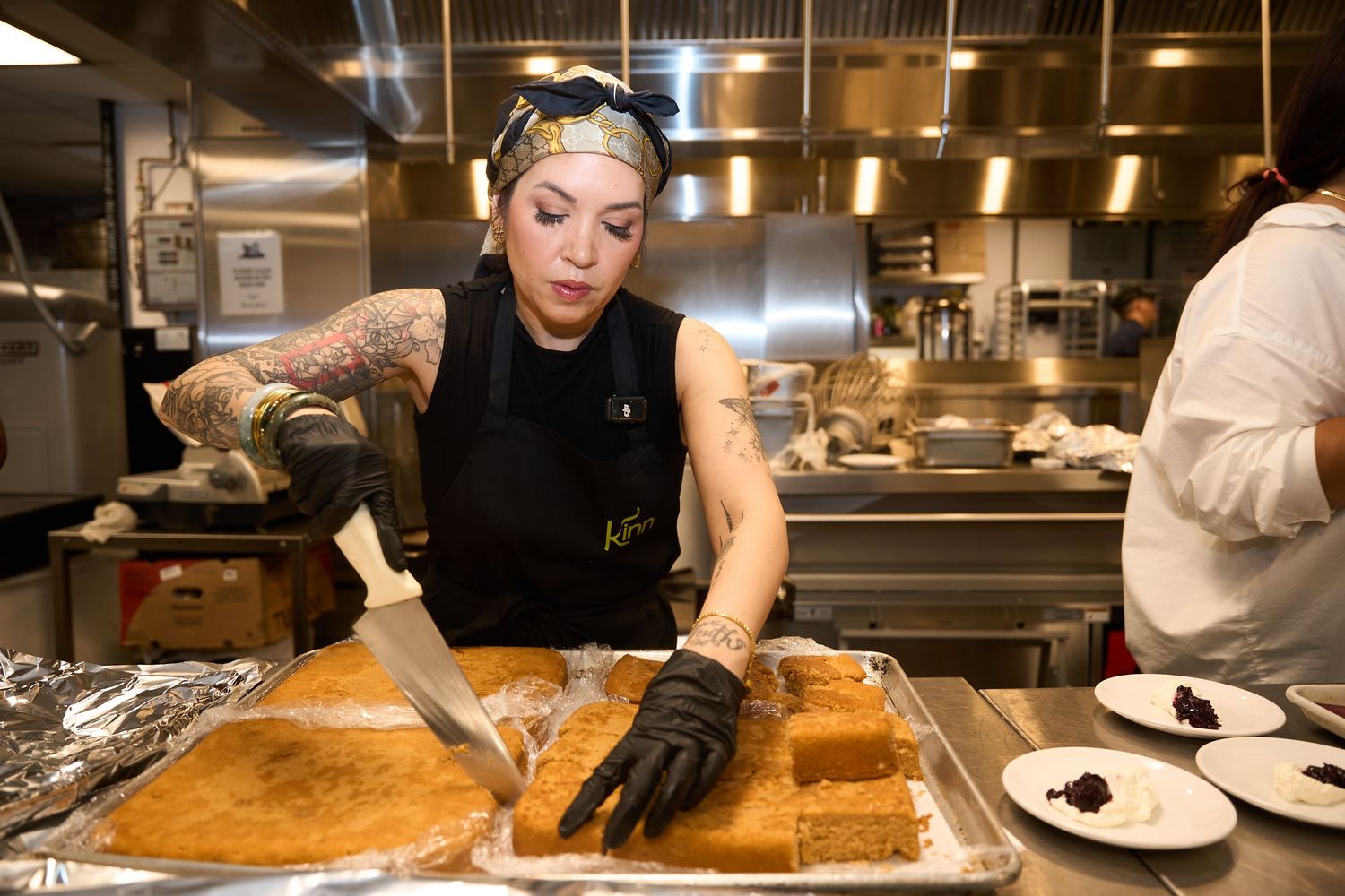 Tattooed chef in a black apron and gloves slicing a golden baked dessert in a metal pan inside a busy commercial kitchen.