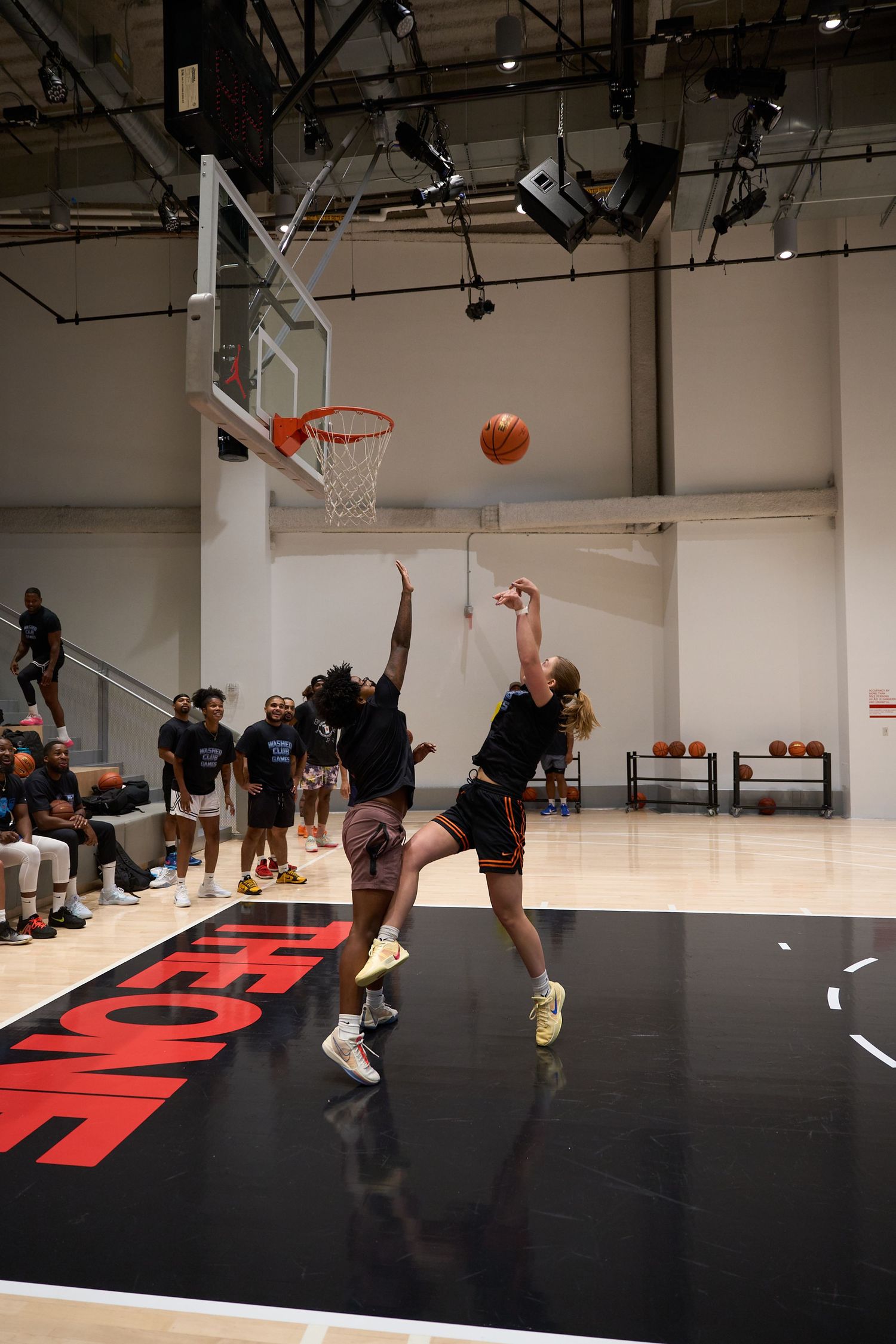 Basketball players practicing dribbling drills on a black court with red markings in an indoor gym facility.