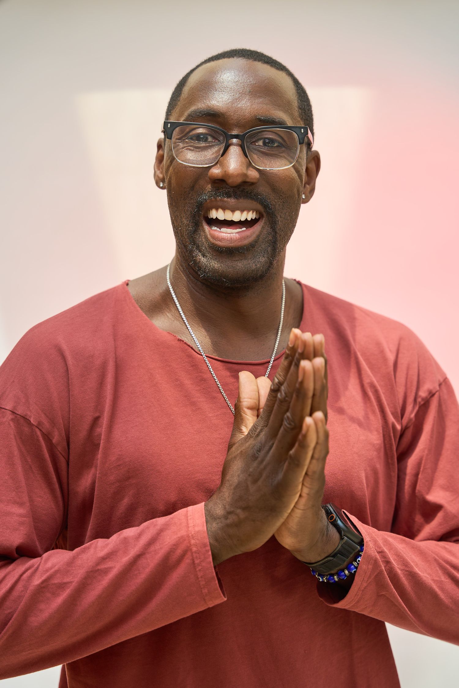 Smiling Black man in red shirt, glasses, and bracelets pressing palms together in a prayer gesture.