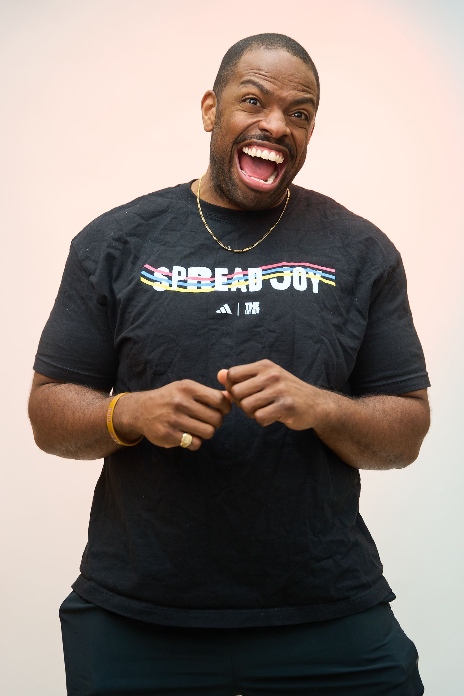 Man in black Adidas Spread Joy t-shirt laughing enthusiastically, wearing gold chain necklace and bracelet.