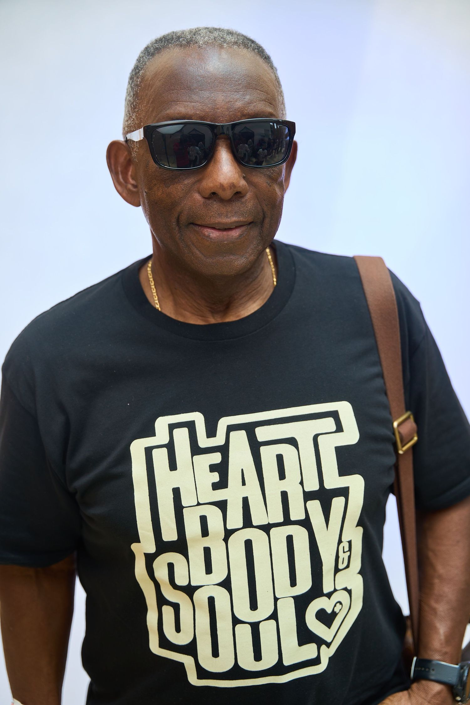 Older man wearing black Heart Body Soul t-shirt and sunglasses with brown shoulder bag strap.