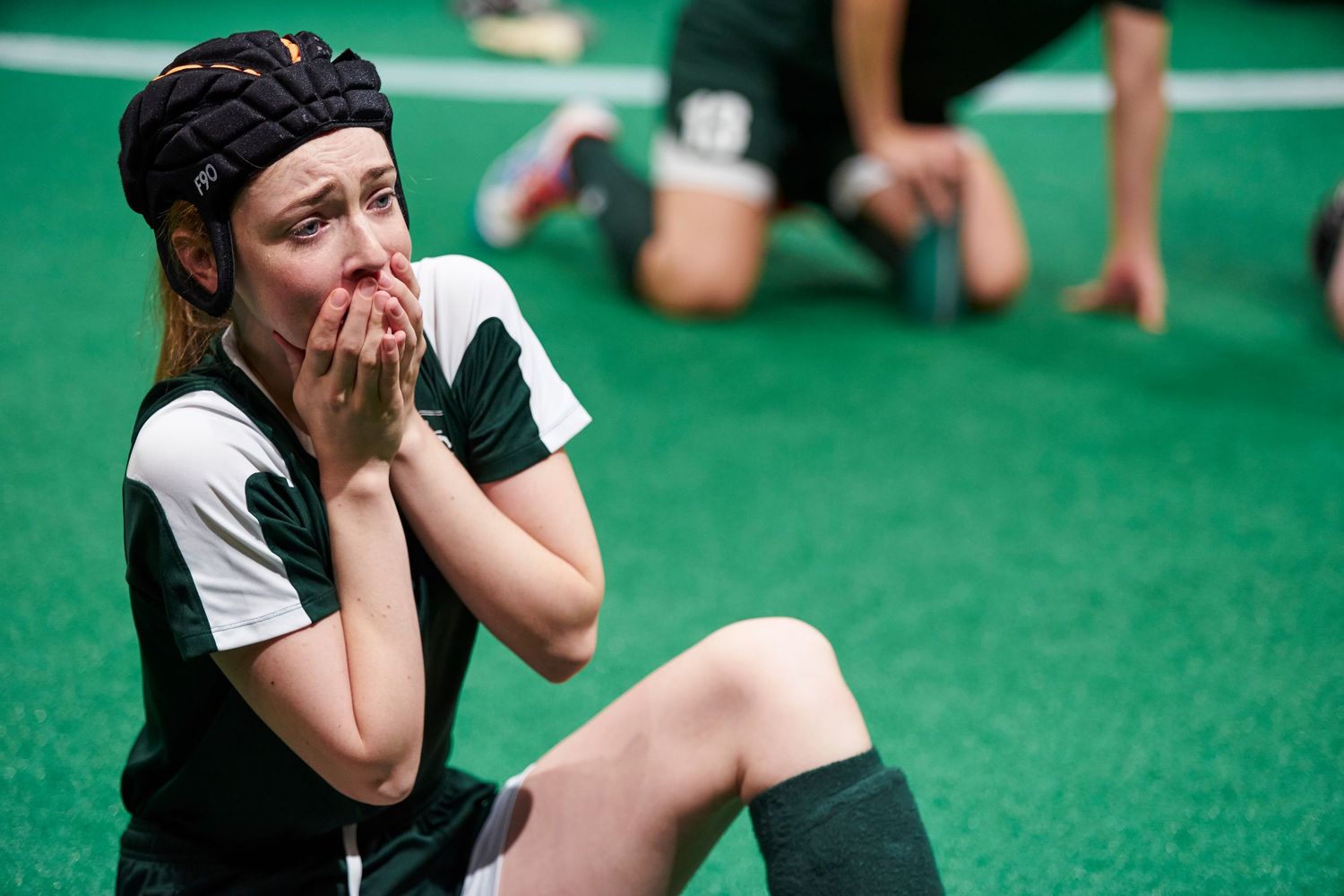 A person sits on artificial turf wearing white and black athletic gear while looking thoughtful with hand on face.