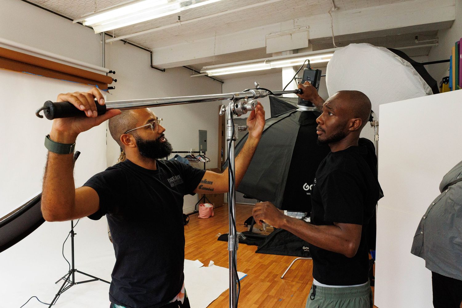 Two musicians in black shirts perform vocals together in a recording studio space with microphones.