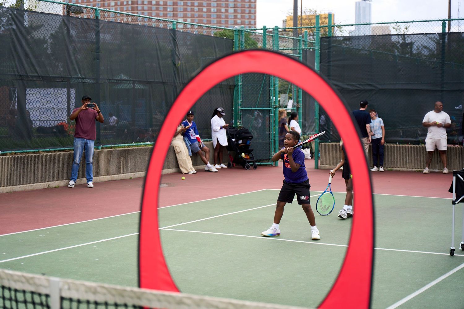 Tennis players practicing serves and volleys on outdoor courts with tall buildings visible in the background.