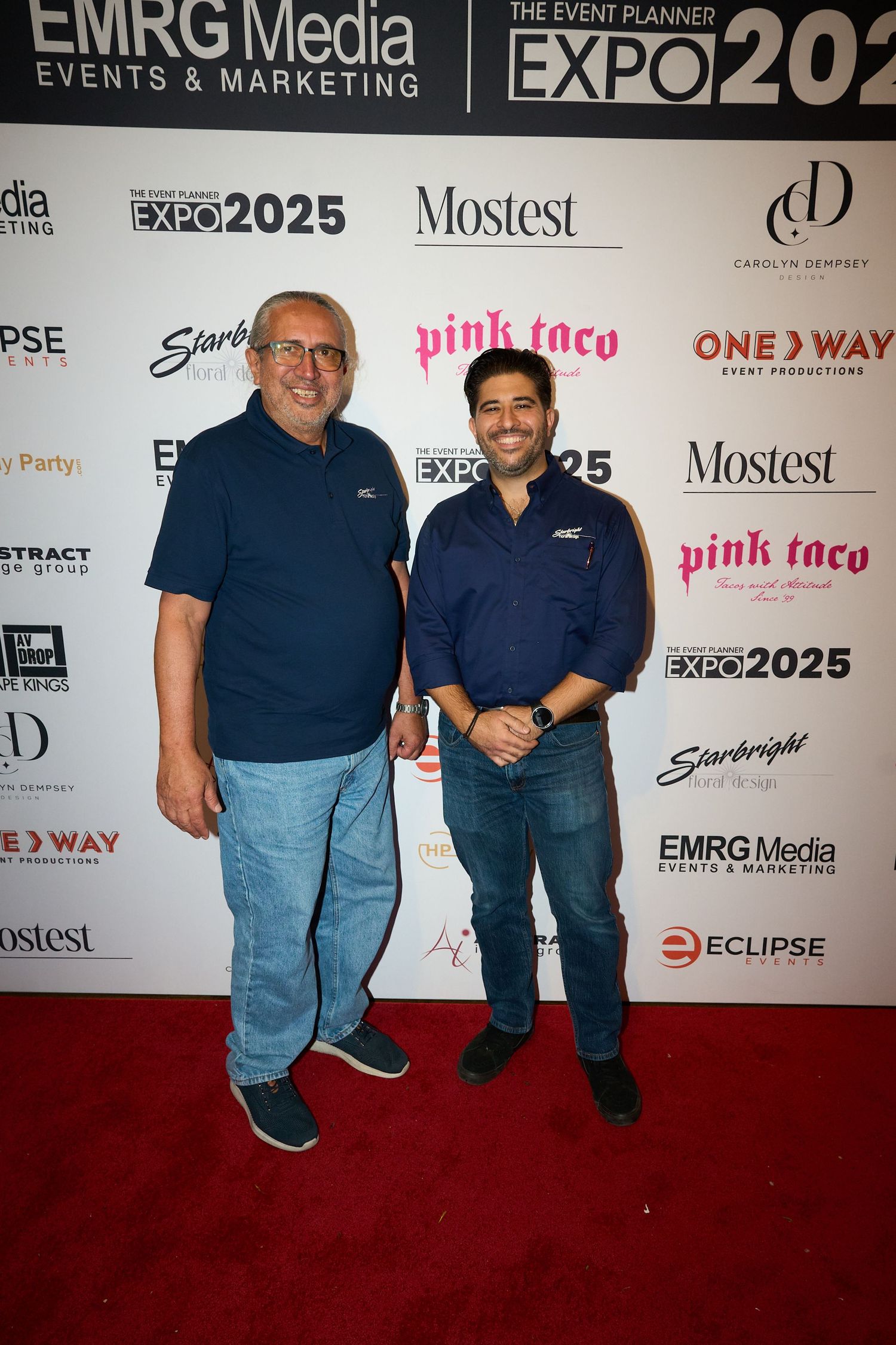 Two people pose together on a red carpet at an event with multiple sponsor logos visible on the backdrop.