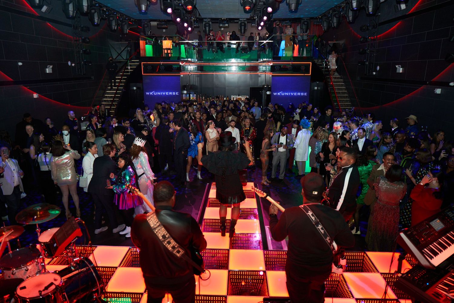 A crowded nightclub dance floor with checkered pattern illuminated by red lighting and colorful flags hanging overhead.
