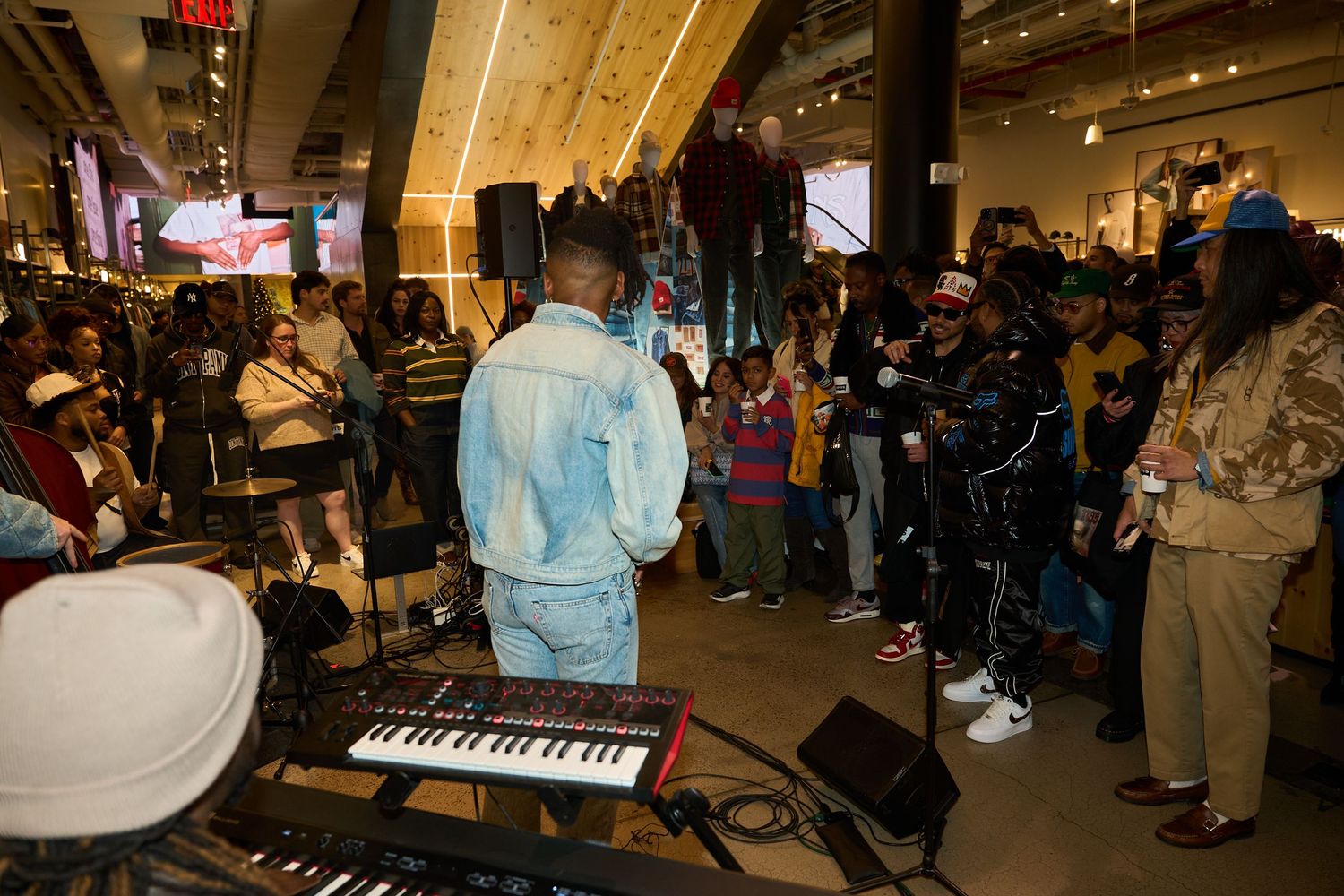 A crowd gathers around performers at an indoor event with musical instruments and dim lighting.