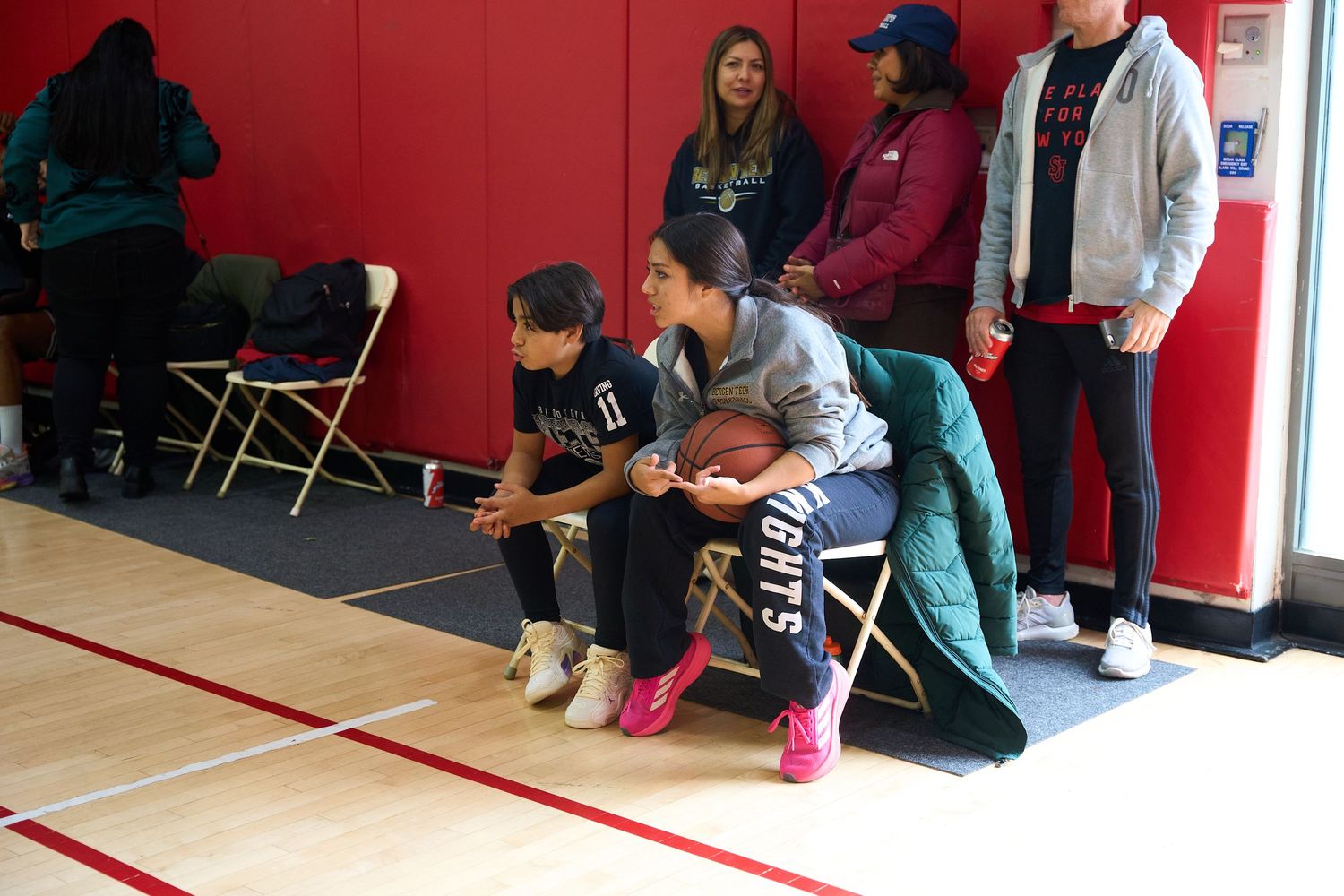 People sit along the sidelines of an indoor basketball court with red walls watching an off-camera game or event.