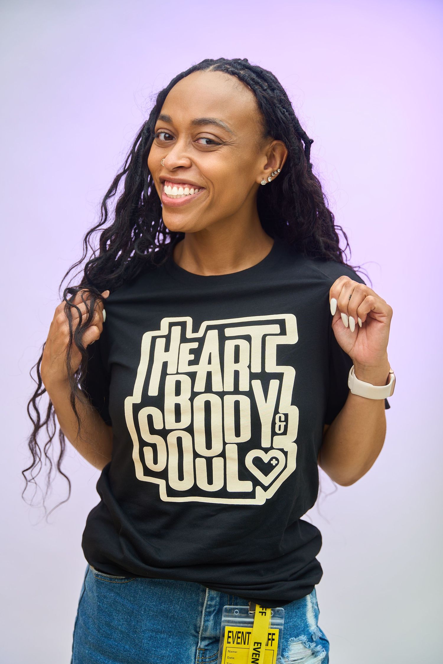 Smiling woman wearing a black Heart Body & Soul graphic t-shirt, with long braided hair, against a purple background.