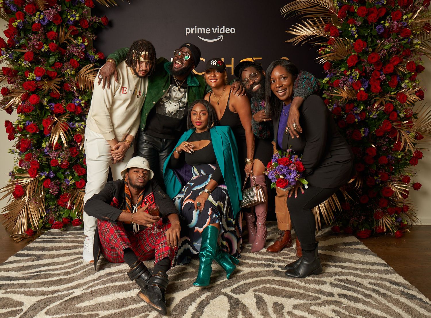 People sitting on a decorative floor pose together in front of a wall covered in vibrant red flowers and greenery.