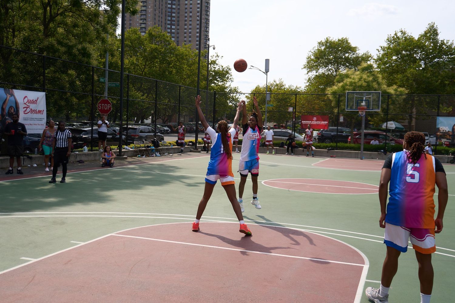 Basketball players compete during a pickup game on an outdoor court in an urban setting with tall buildings in background.