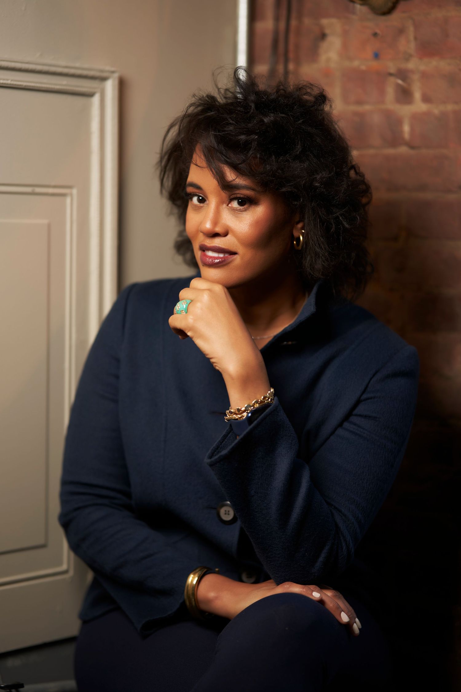Professional portrait of someone wearing a navy blazer seated against a brick wall background with elegant jewelry and makeup.