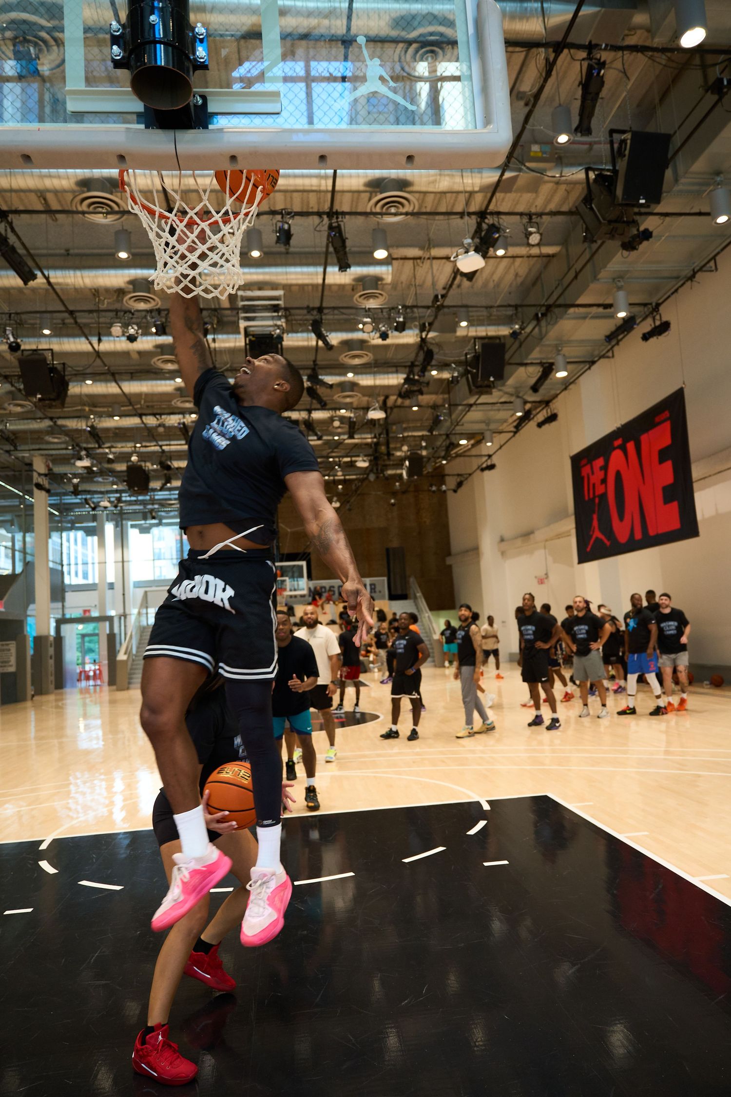 Basketball players practicing drills on a well-lit indoor court with high ceilings and large windows.